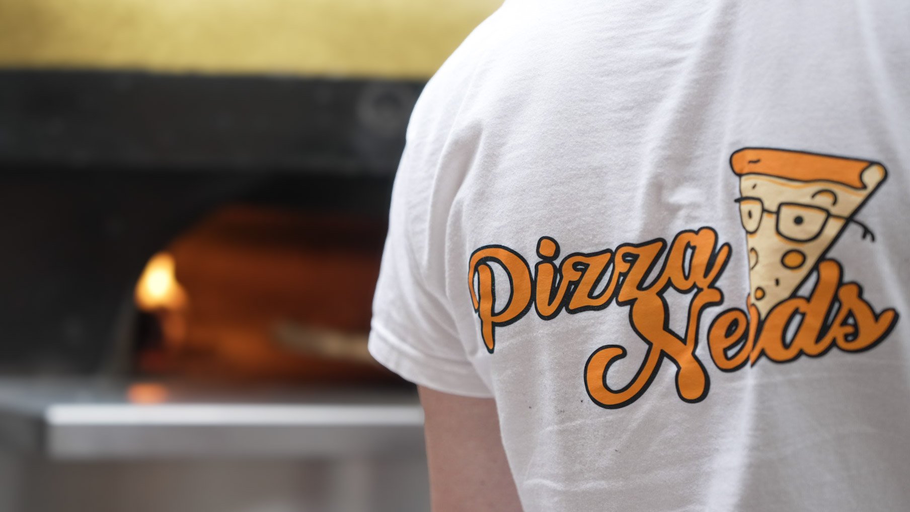 Close-up of a person wearing a white T-shirt with a logo for 'Pizza Seeds,' featuring an animated slice of pizza wearing glasses and a chef's hat, with a pizza oven in the background.