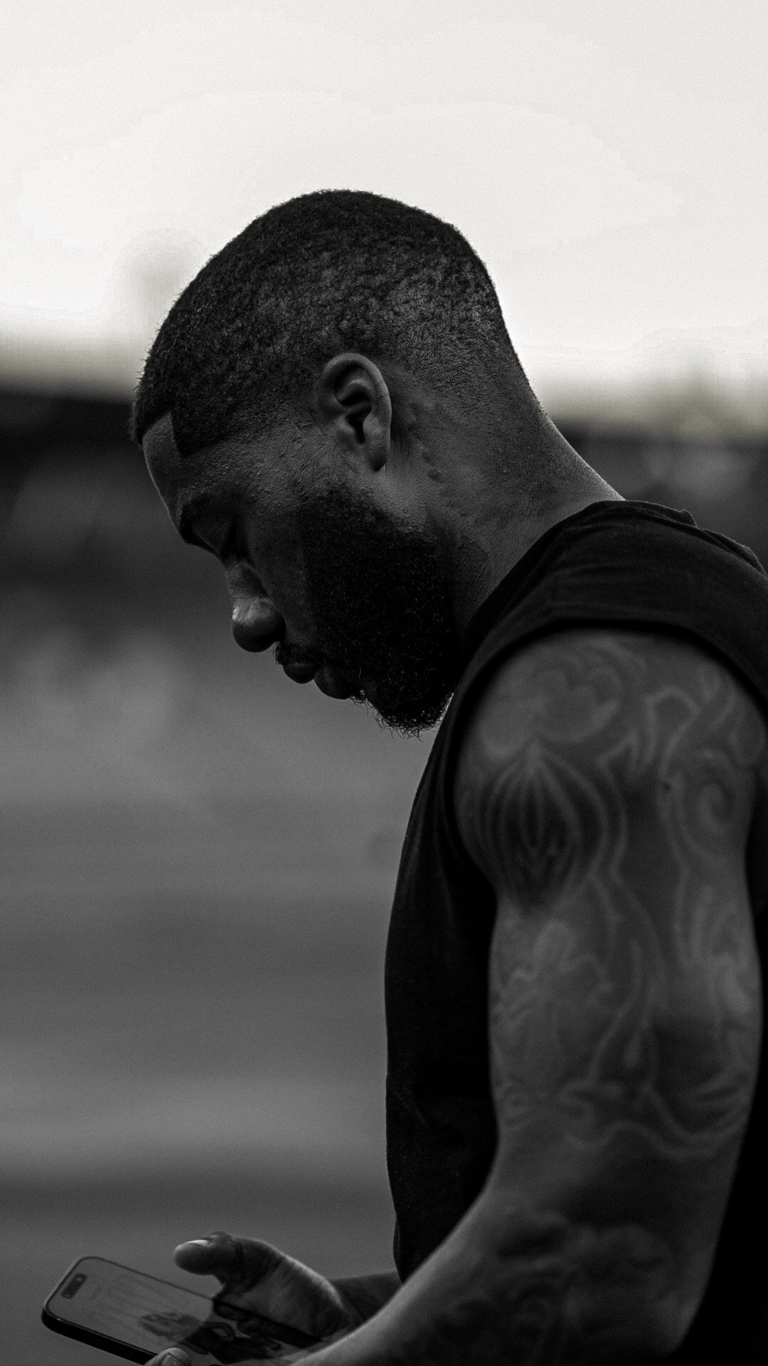 A young man with a beard and tattoos on his upper arm, looking down at a smartphone, in black and white.