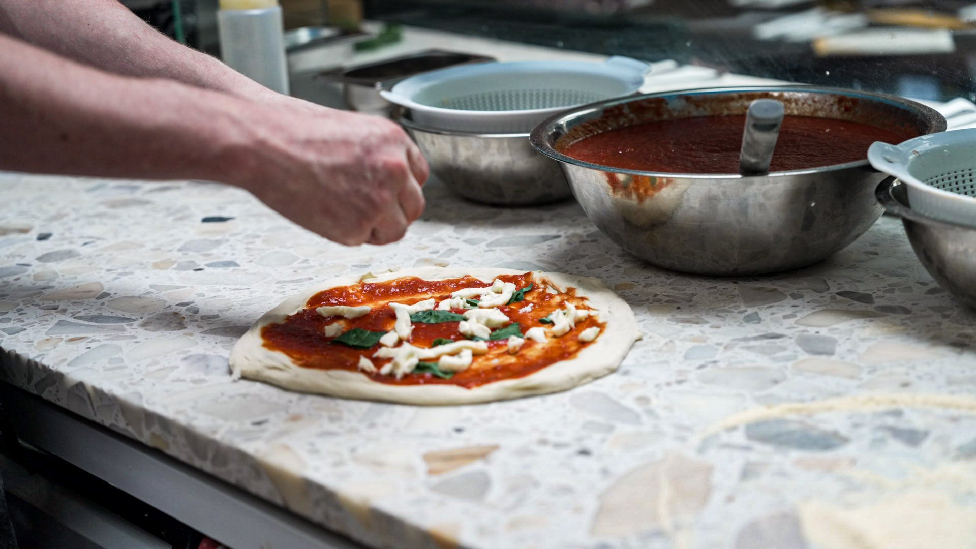 Person preparing a pizza with tomato sauce, cheese, and basil on a marble countertop with various bowls of ingredients in the background.