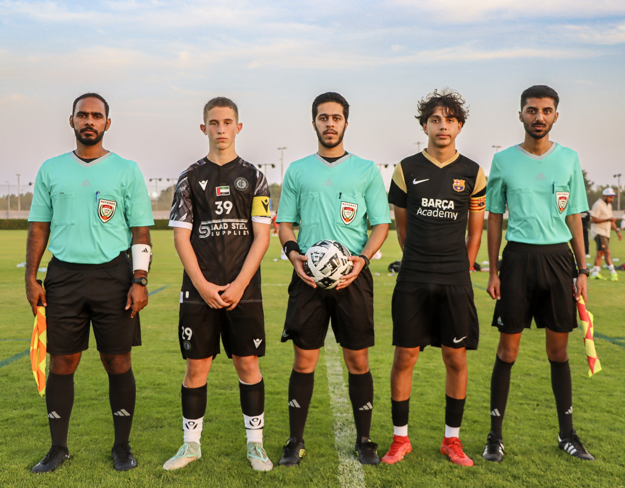 Five soccer players standing on the field before a match, with two referees flanking them, holding a soccer ball. The players are wearing jerseys representing different teams, with a mix of black and light turquoise shirts. The referees are wearing t