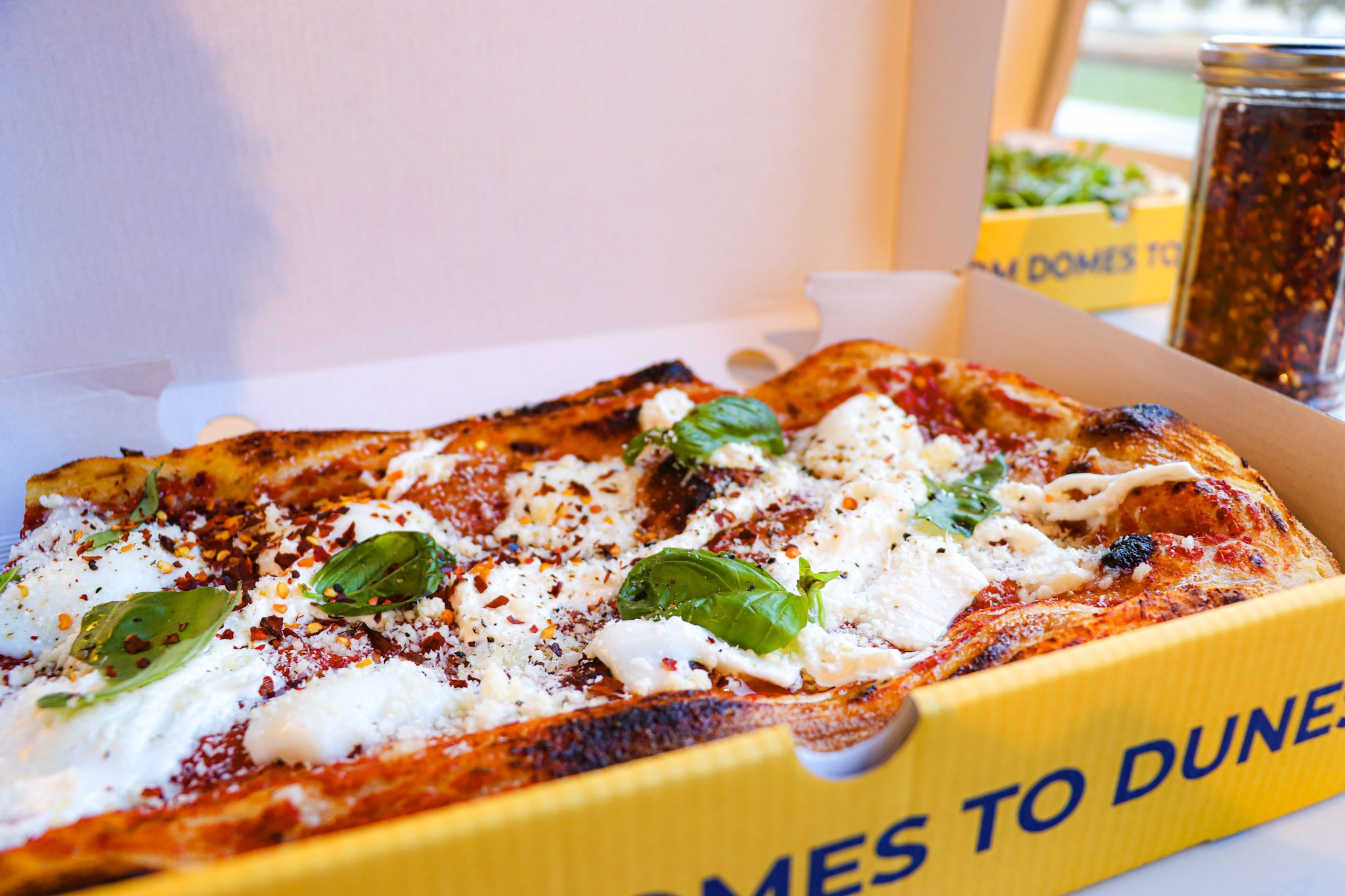 A pizza in a pizza box with cheese, tomato sauce, basil, and spices. A container of red pepper flakes is visible in the background.