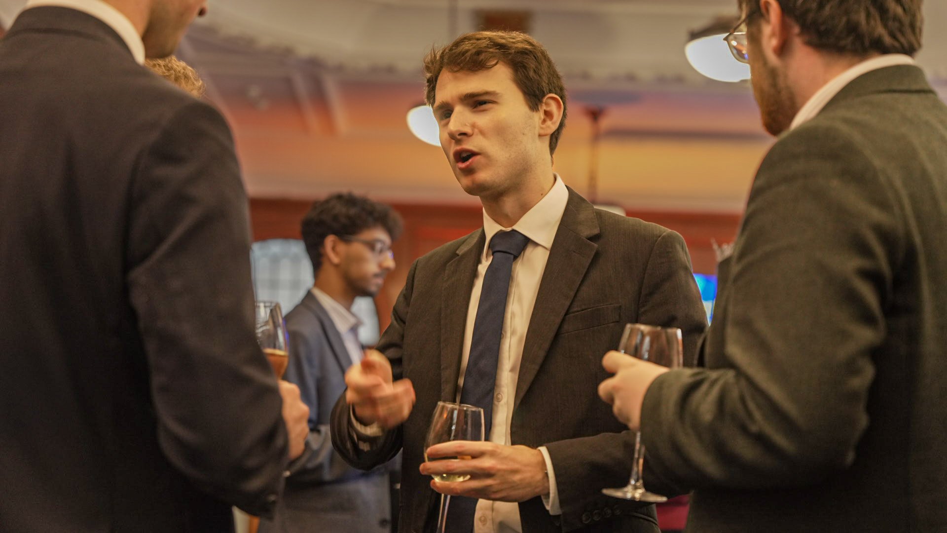A man in a gray suit and blue tie speaking to two other men in suits at a social event, holding a glass of white wine, with other guests in the background.