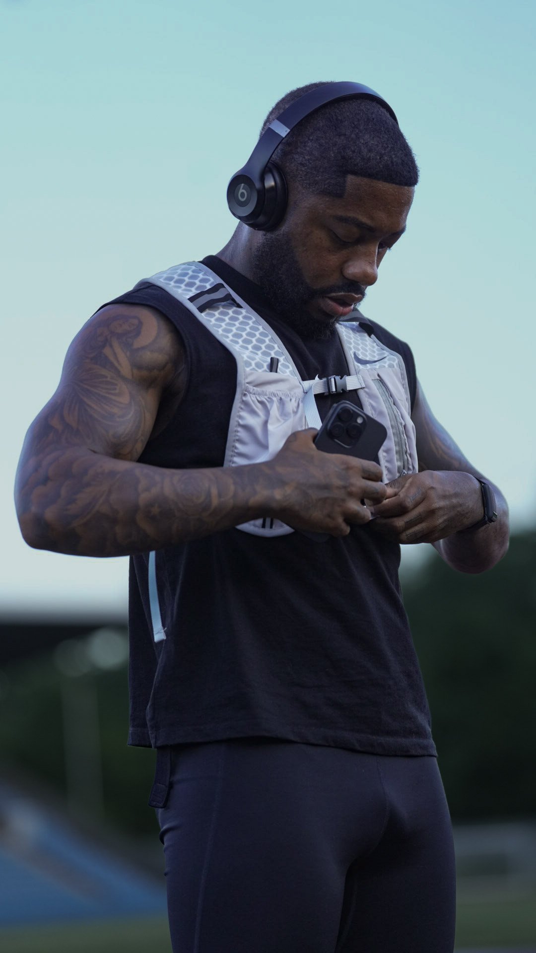 Man with tattoos wearing a black sleeveless shirt and gray running vest, holding a smartphone with headphones on, outdoors during evening or early morning.