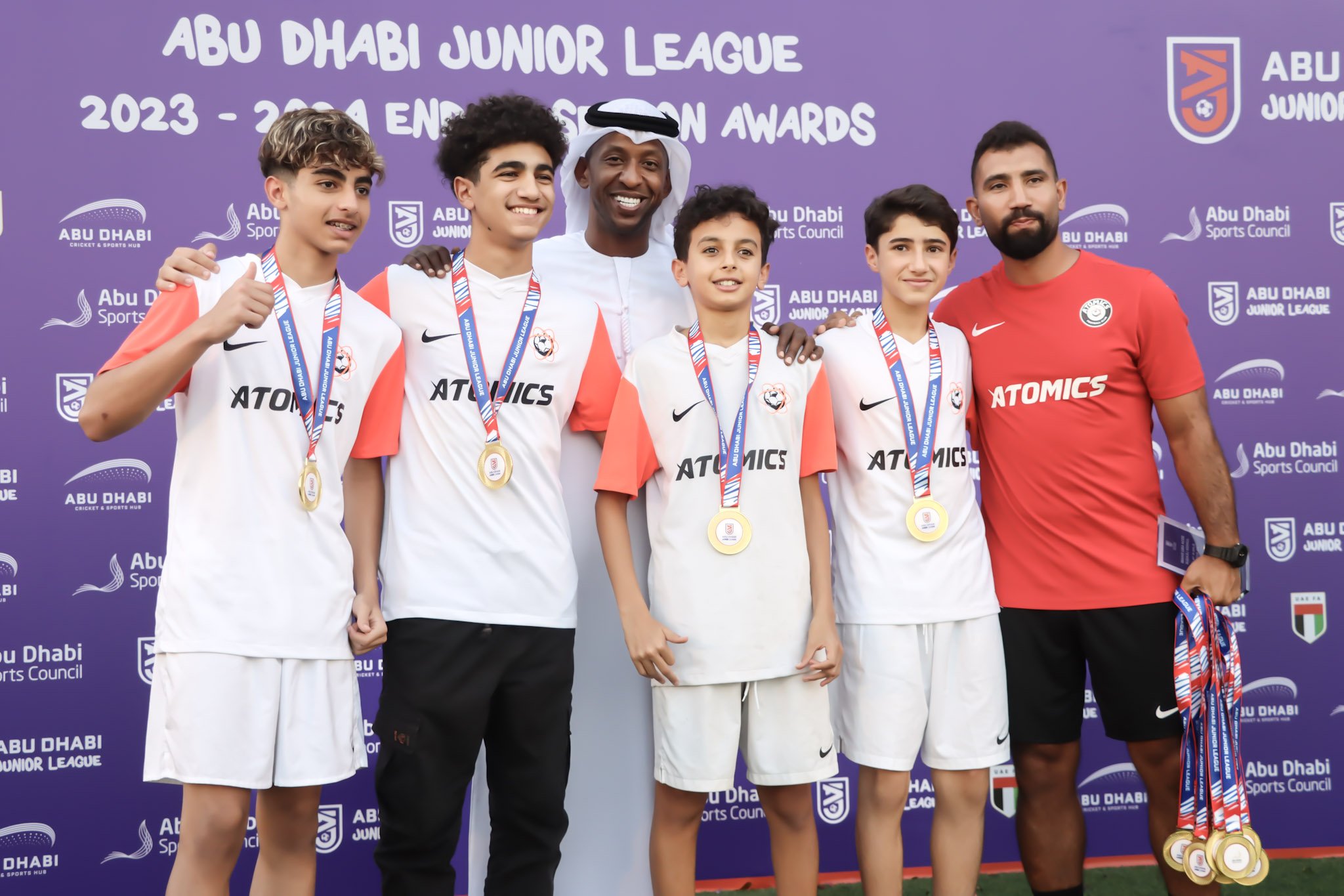 Group of five people, four young boys and one adult man, standing on a podium with medals around their necks, smiling after winning a sports event. They are in front of a purple backdrop with text and logos related to the Abu Dhabi Junior League awar