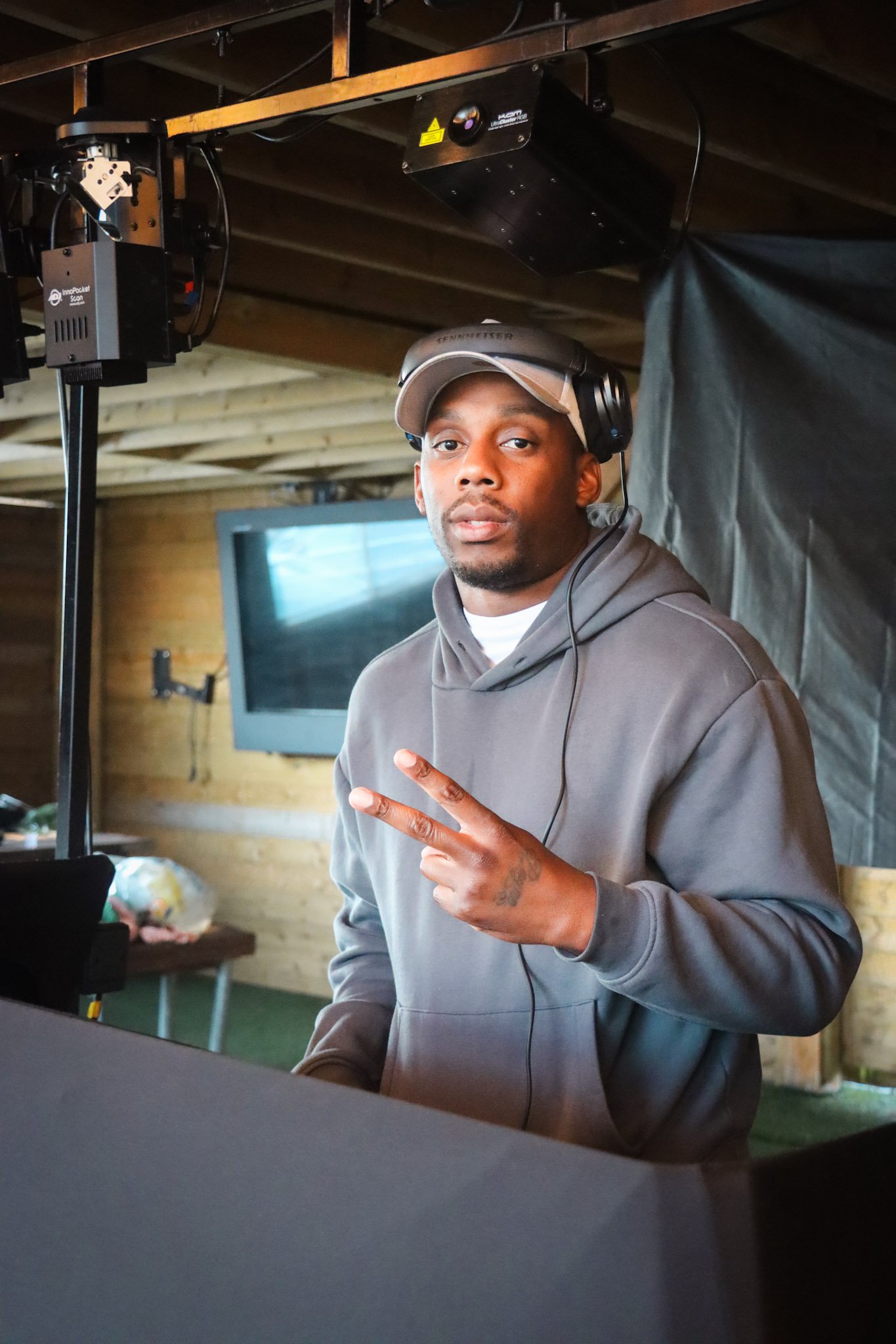 A young man wearing headphones and a gray hoodie making a peace sign with his fingers in a rustic indoor setting with wood walls, a television, and equipment.