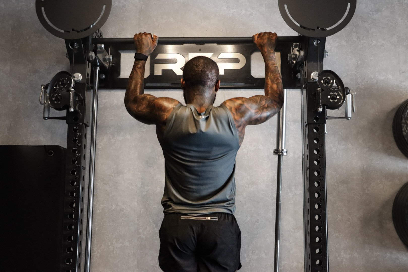 A muscular man with tattoos performing pull-ups on a black pull-up bar in a gym, with weights on the wall on the right side.