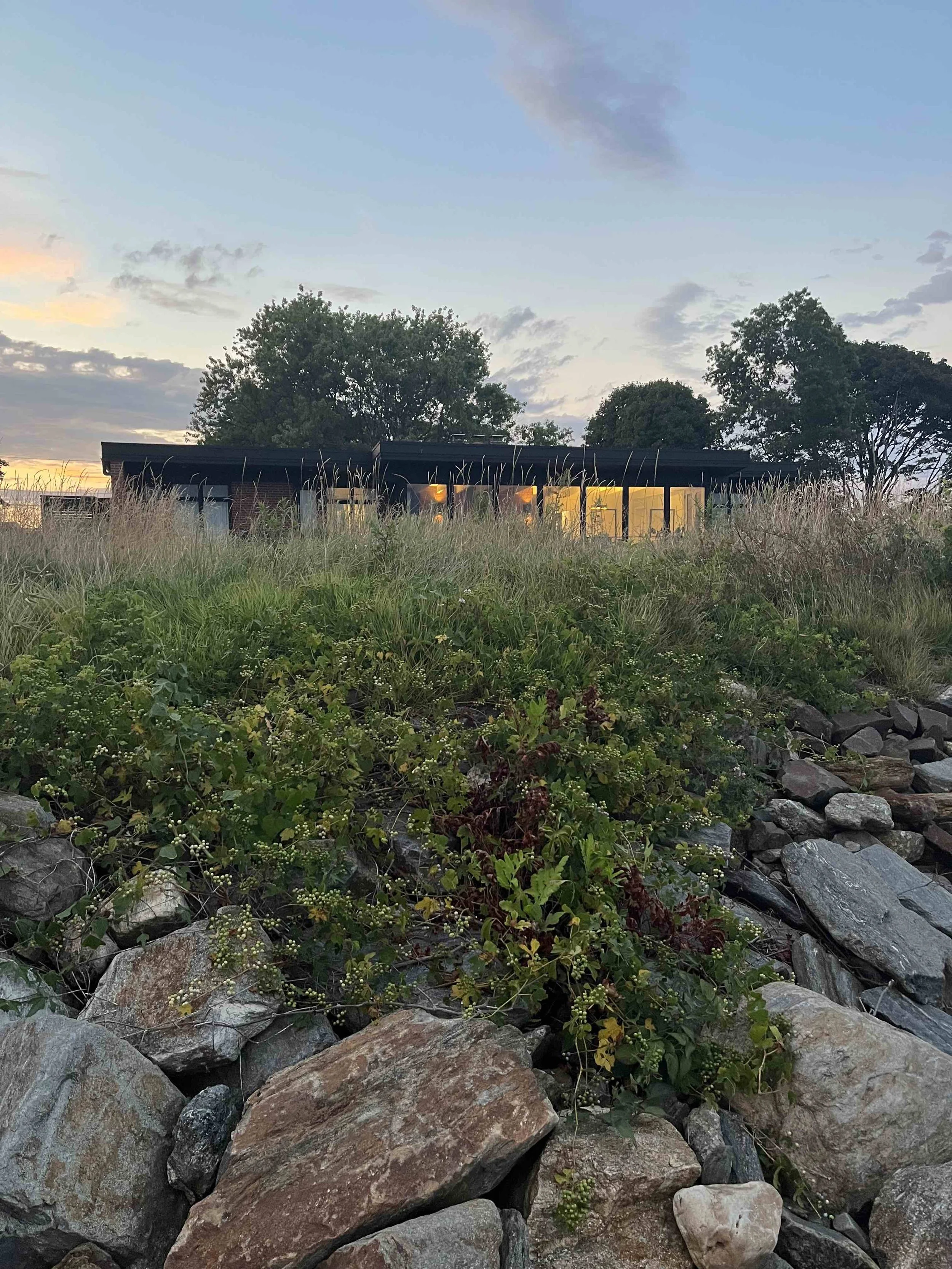 photo of black roofline of lordship house as seen from below a rocky bluff, the windows softly glow yellow, an early evening sky above