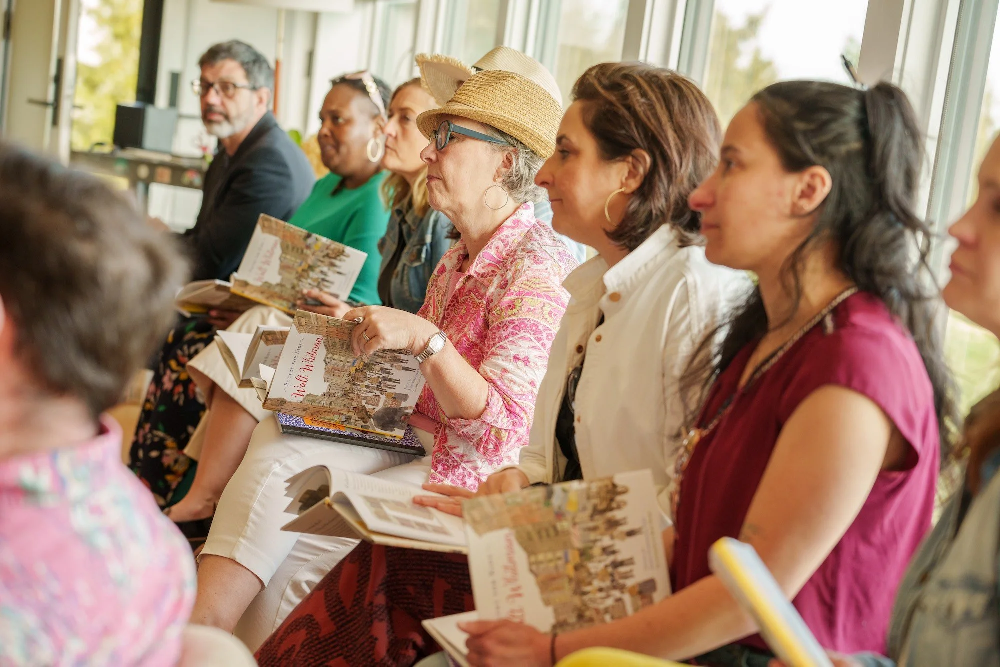 Photo of community members, seated on a bench in front of light-filled windows, hold copies of "Walt Whitman: Poetry for Kids" by Dr. Karen Karbiener. 