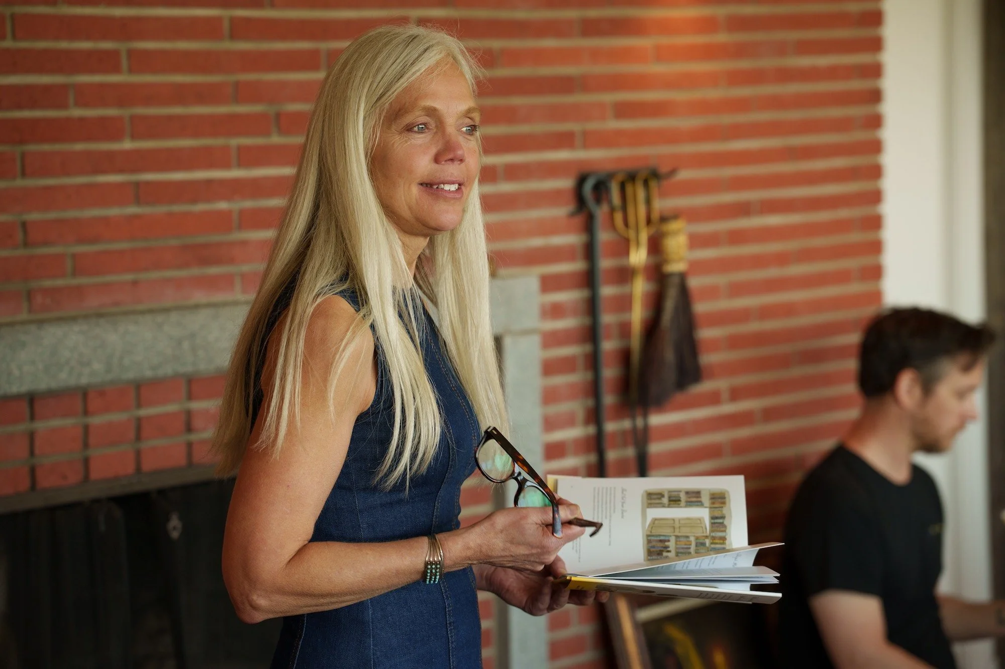 photo of Dr. Karen Karbiener who holds glasses and an open book in her hands. She is smiling and looking off-camera at audience. She is framed by a red brick wall.