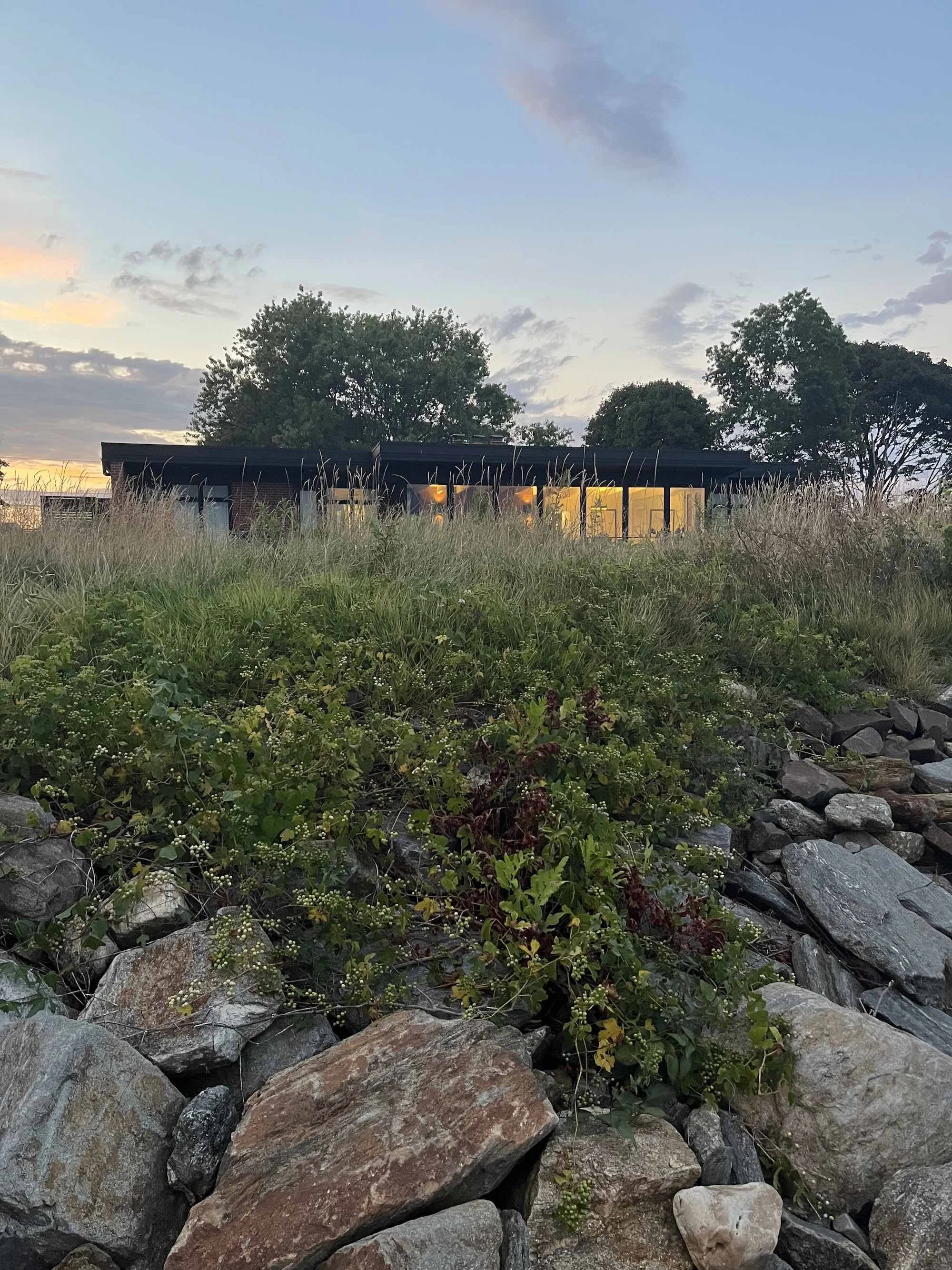 photo of black roofline of lordship house as seen from below a rocky bluff, the windows softly glow yellow, an early evening sky above
