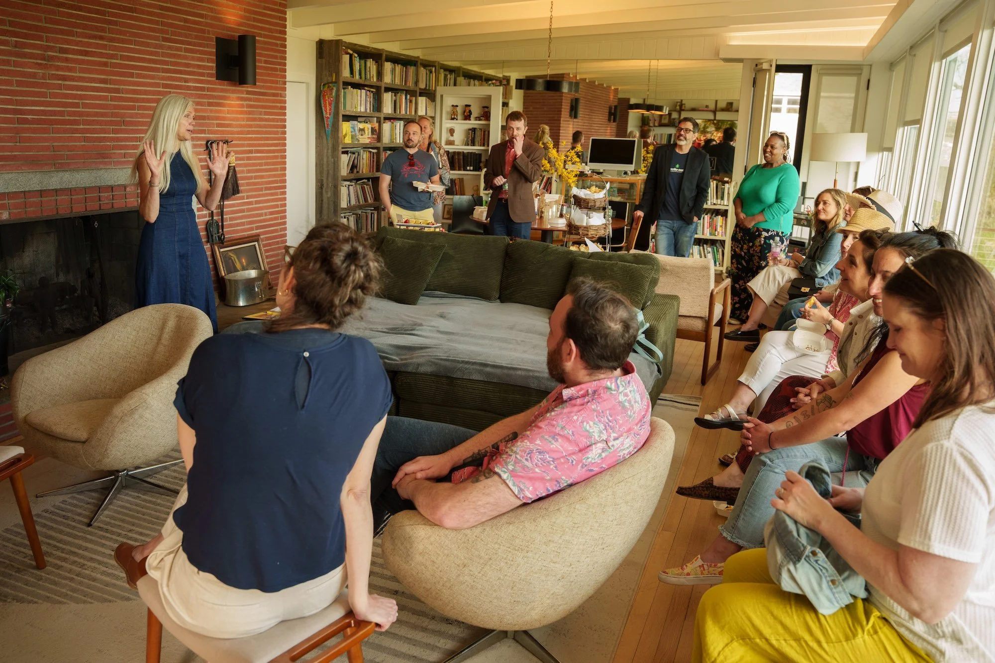 photo of Dr. Karen Karbiener who stands in front of a brick hearth, gesturing with her hands in front of an audience of a dozen people, seated along a window bank and in various chairs around the lordship house living room.