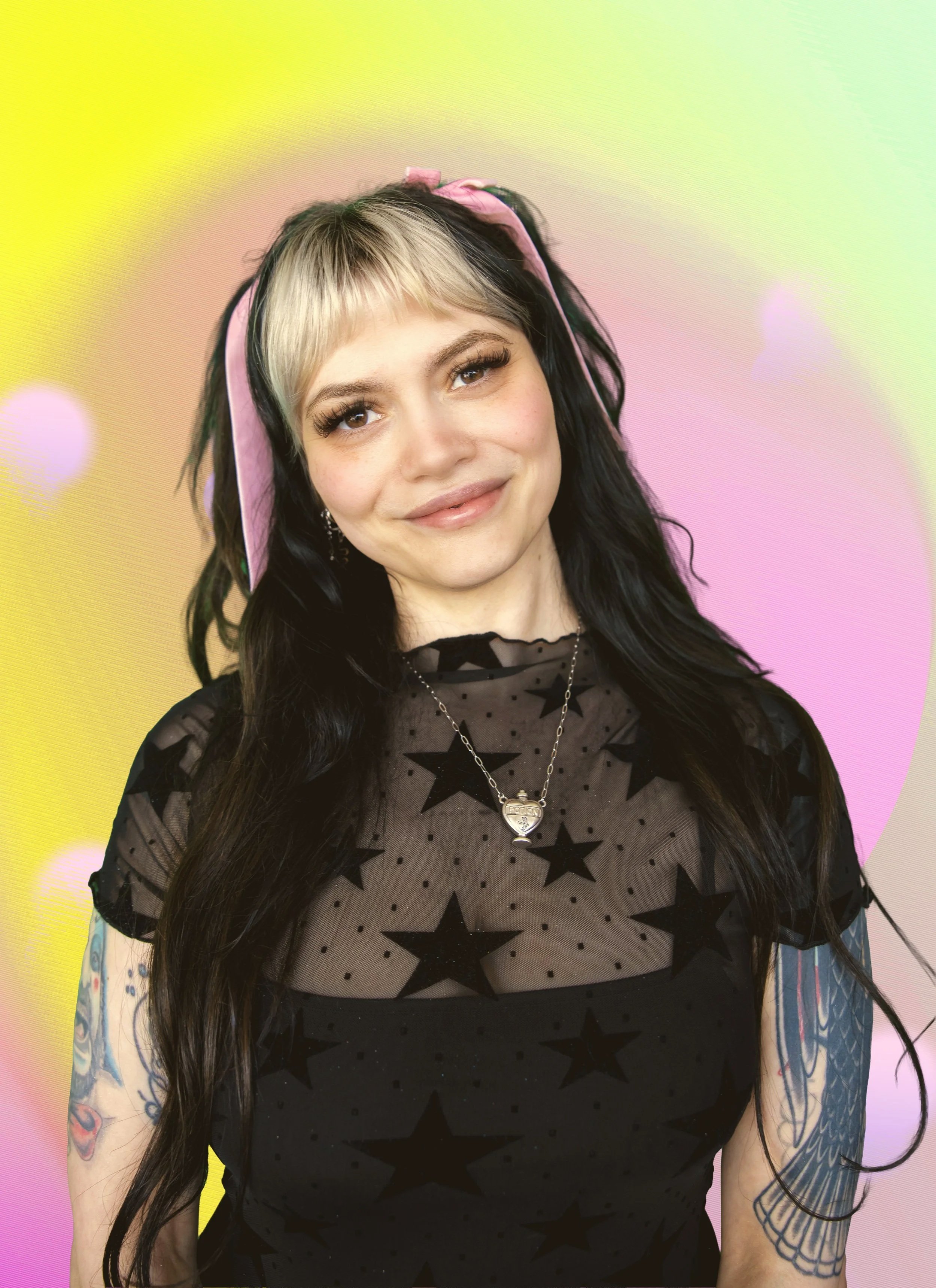 A young woman with blonde and black hair smiling, posing with her hand resting on her neck, in front of a pastel-colored background.