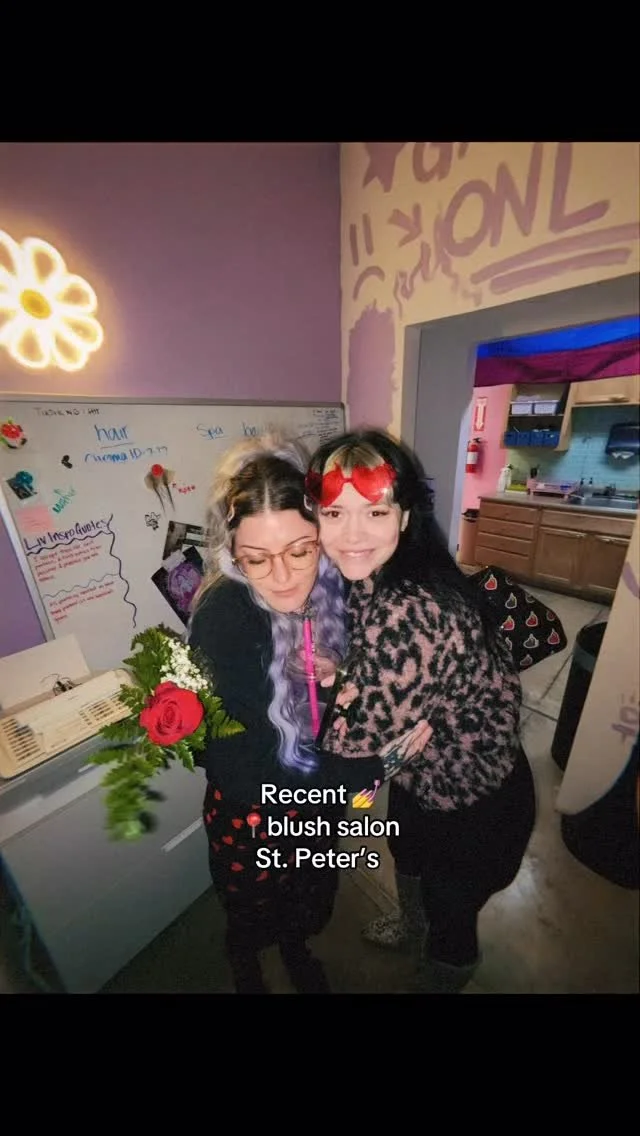 Two women at a salon, one holding a bouquet of flowers, smiling for the camera with text overlay stating "Recent blush salon St. Peter's." The salon has a purple wall, a whiteboard with writing and decorations, and a doorway revealing a kitchen area.
