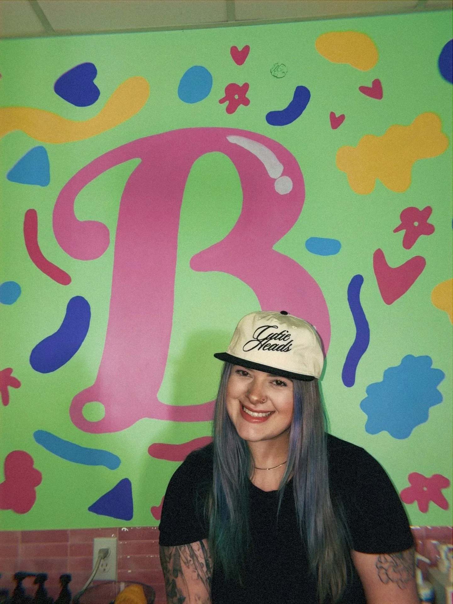 A young woman with multicolored hair and tattooed arms smiling at the camera, wearing a white cap with black trim and text reading 'Gatie Heads,' against a bright green wall decorated with large pink number three and various colorful abstract shapes.