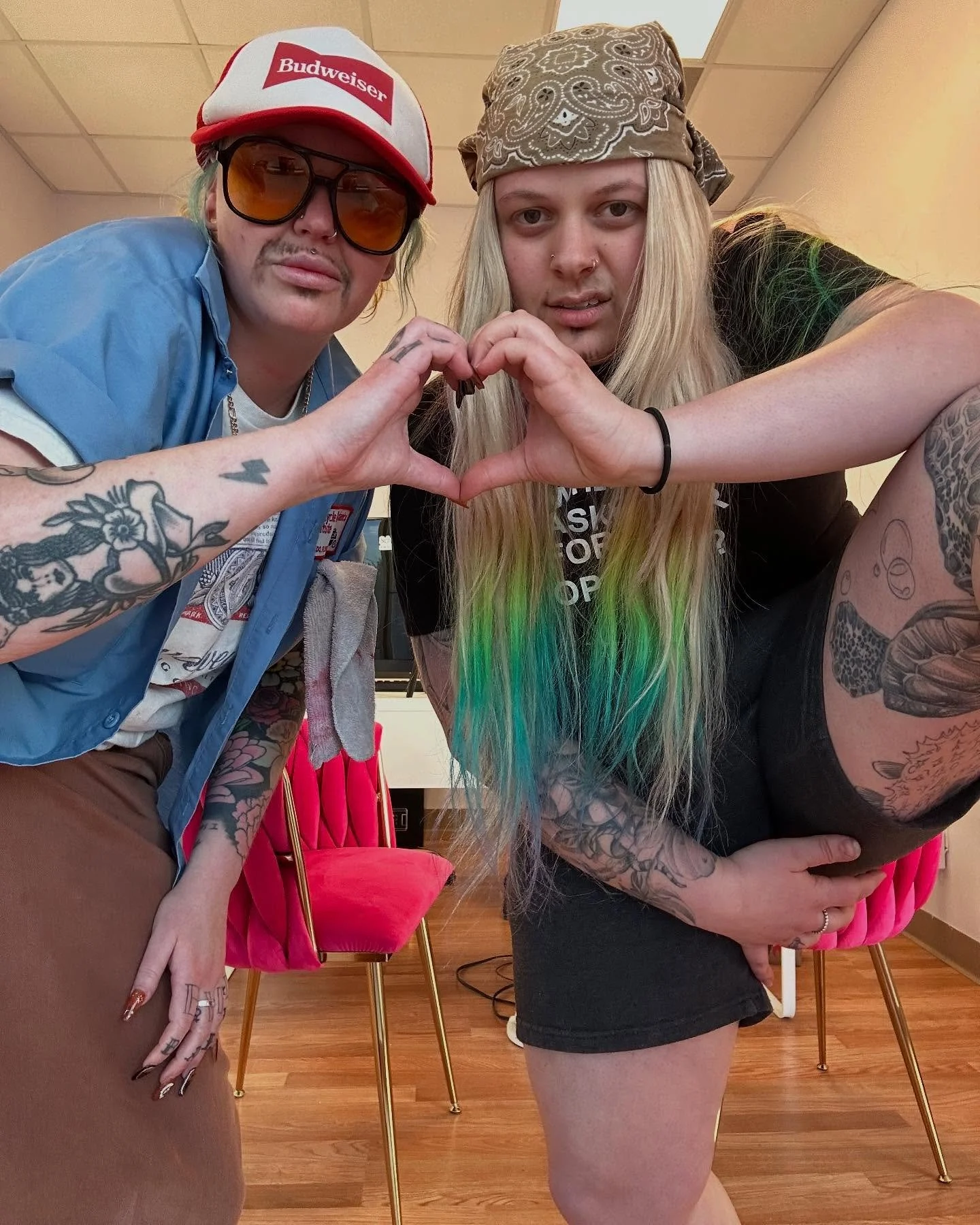 Two women with tattoos forming a heart shape with their hands, posing in an indoor setting with pink chairs and wooden flooring.