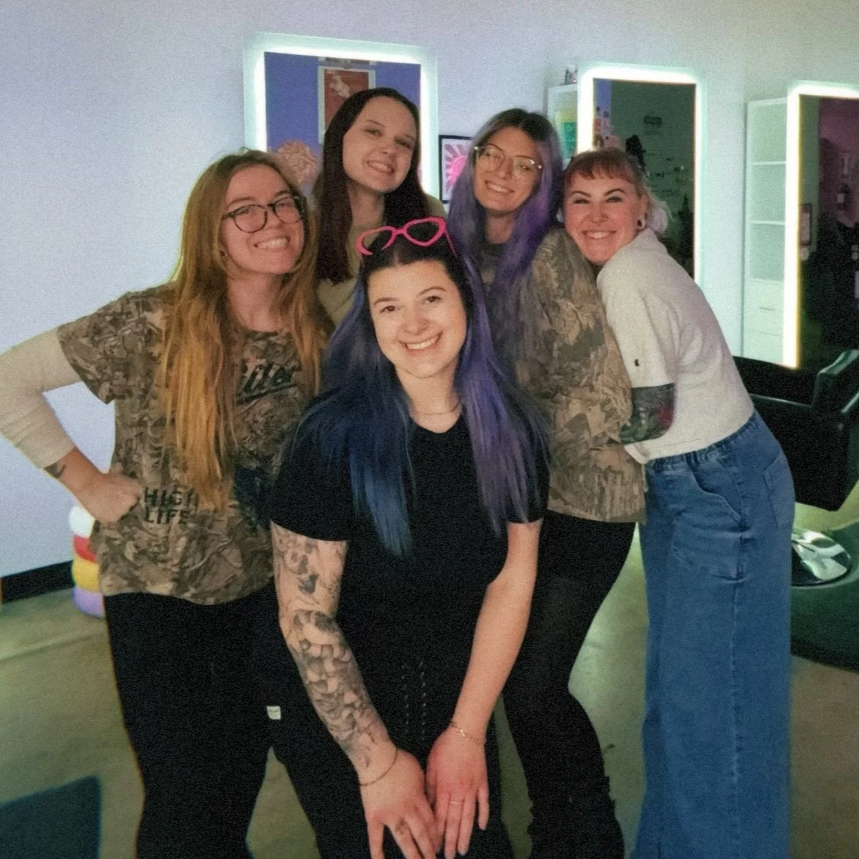 Group of five young women smiling and posing together indoors, with some wearing glasses and colorful hairstyles.