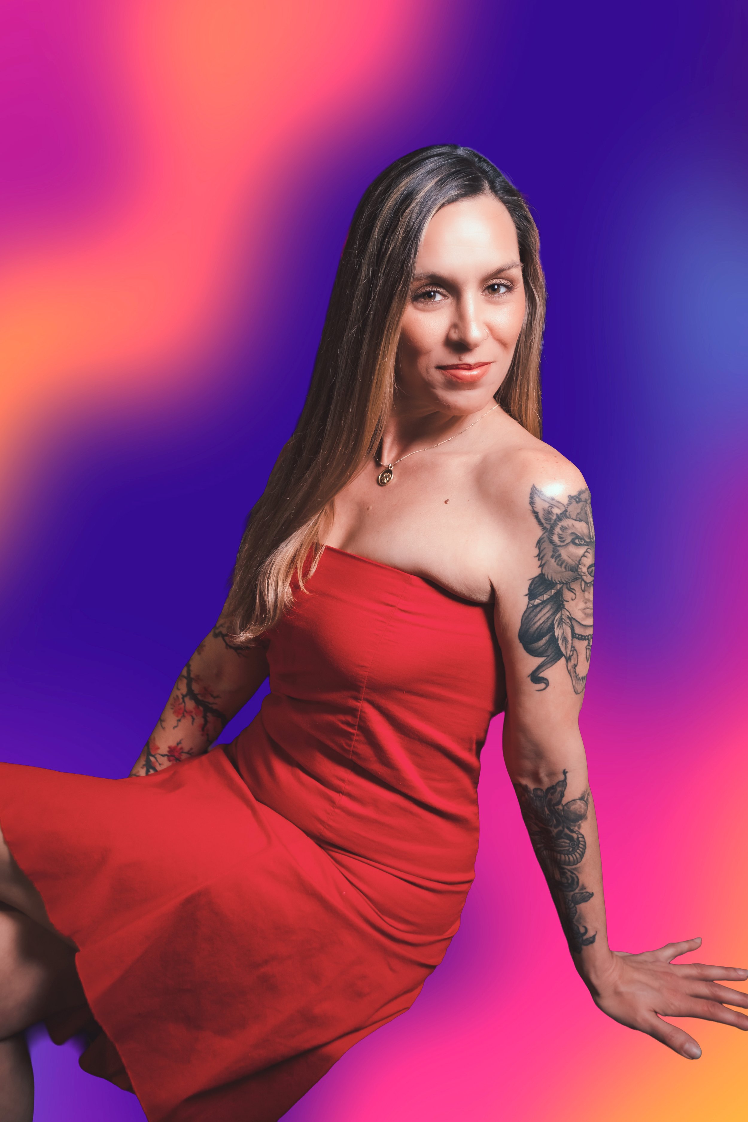 A woman with long blonde hair and tattoos on her left arm poses in front of a pink leopard print background with neon lights. She wears a strapless red dress and looks at the camera with a slight smile.