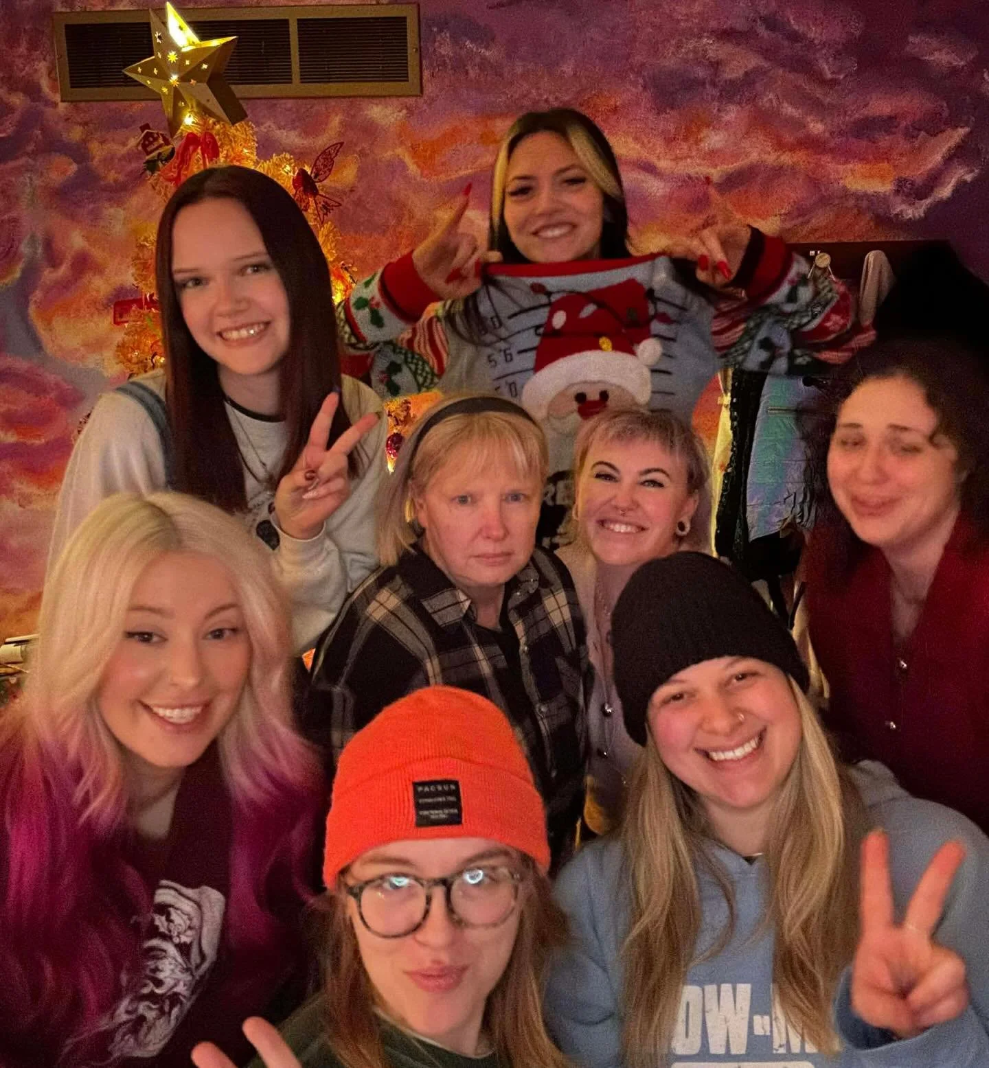 Group of nine women smiling and posing for a photo in front of a colorful, cloudy background with a decorated Christmas tree.