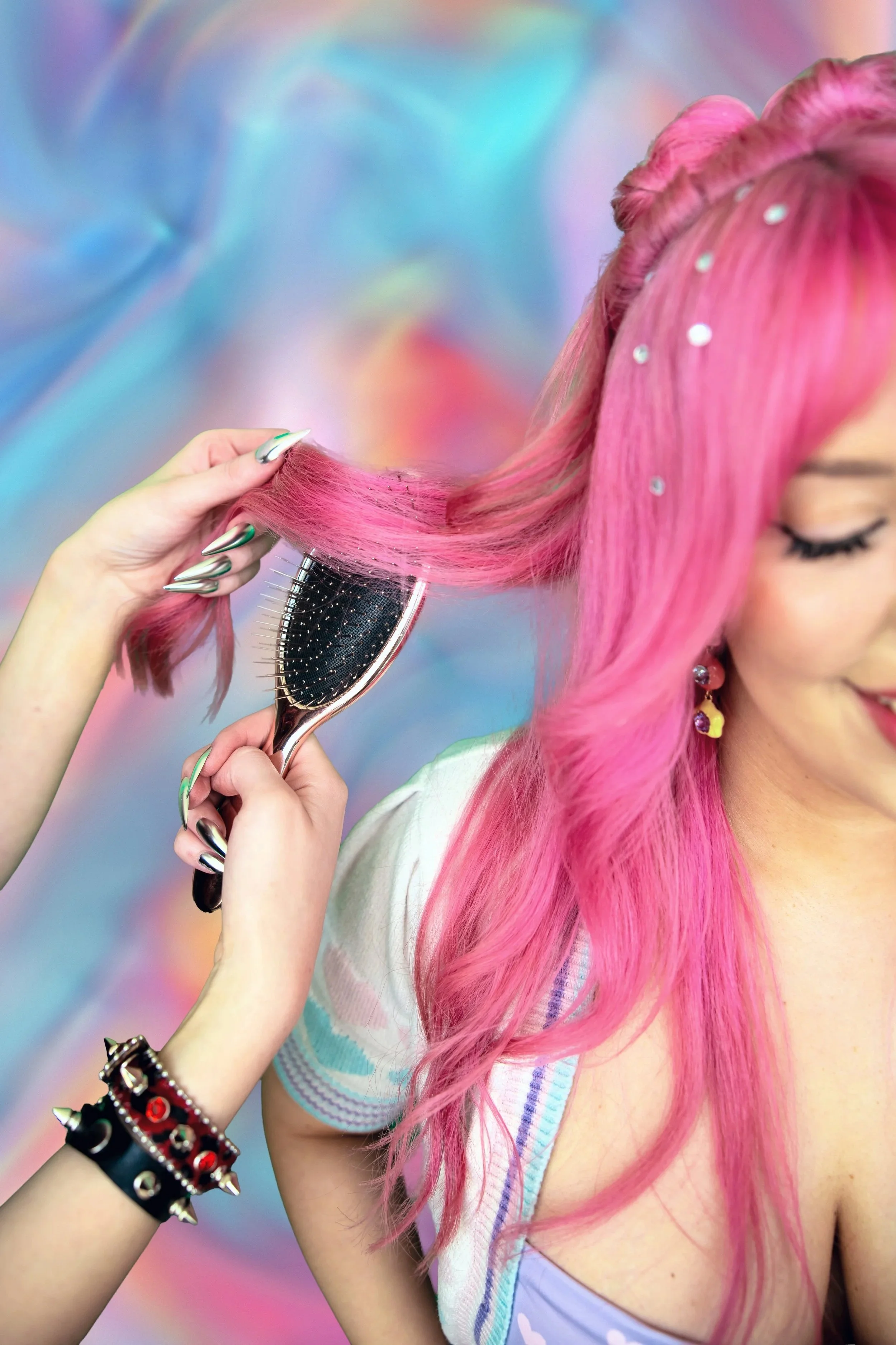 A woman with curly pink hair, topless, with a row of flowers around her waist and her arms raised above her head against a plain background.