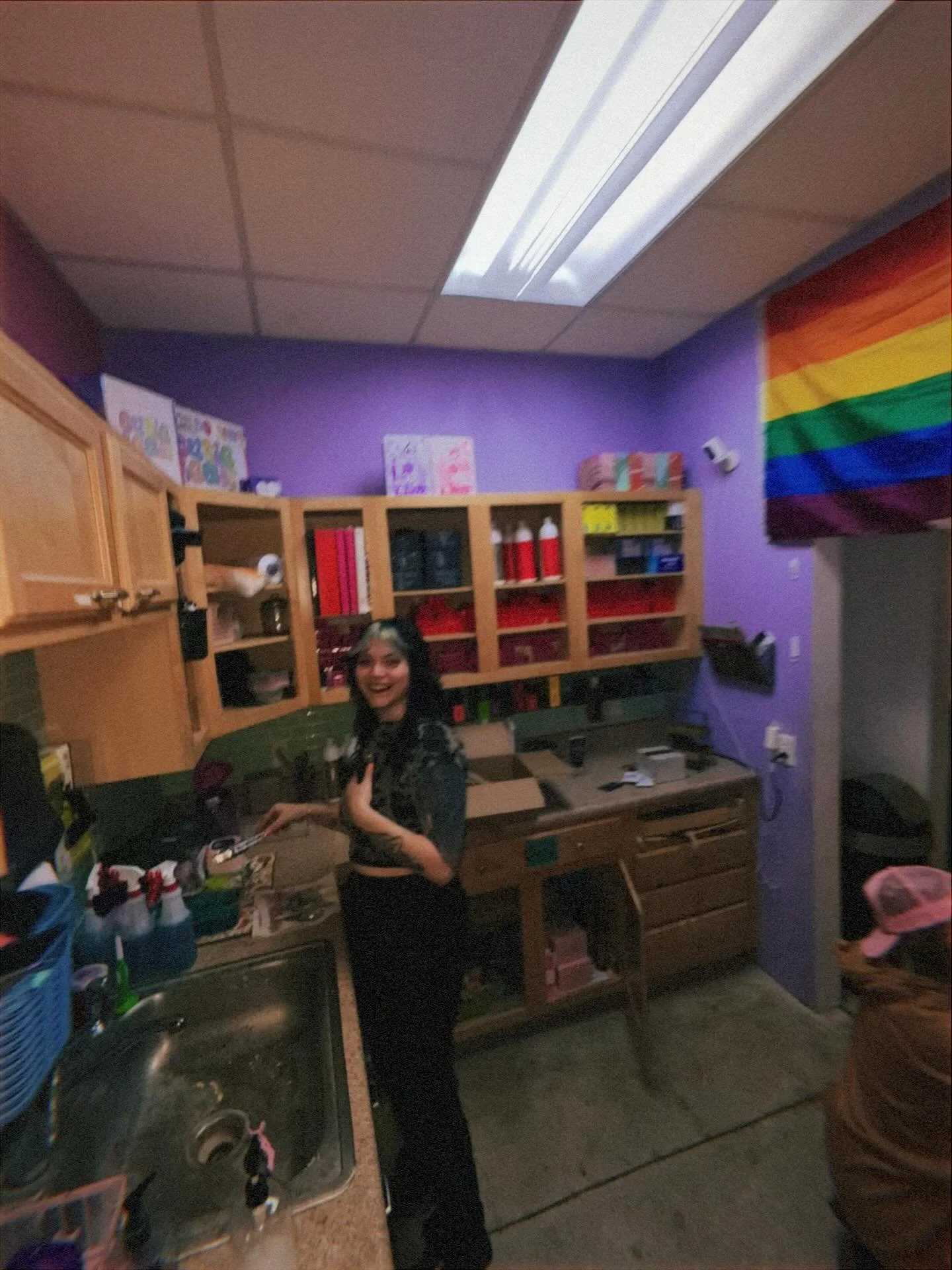 A woman with dark hair smiling in a colorful, cluttered kitchen with a purple wall, rainbow pride flag, and open cabinets filled with red, yellow, and black supplies.