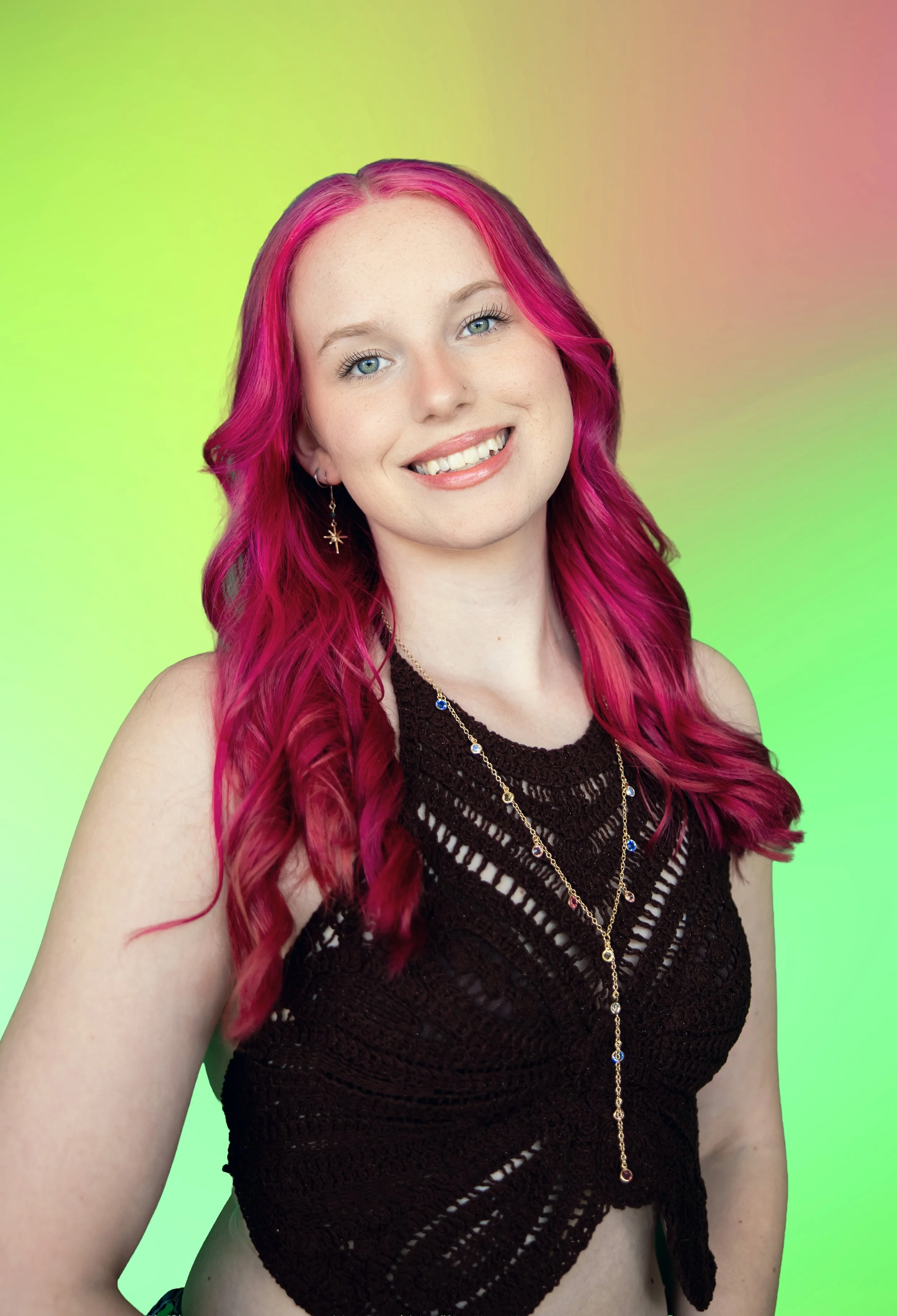 Young woman with long red hair smiling at camera, wearing a green top and layered necklaces, standing in front of a colorful abstract background.