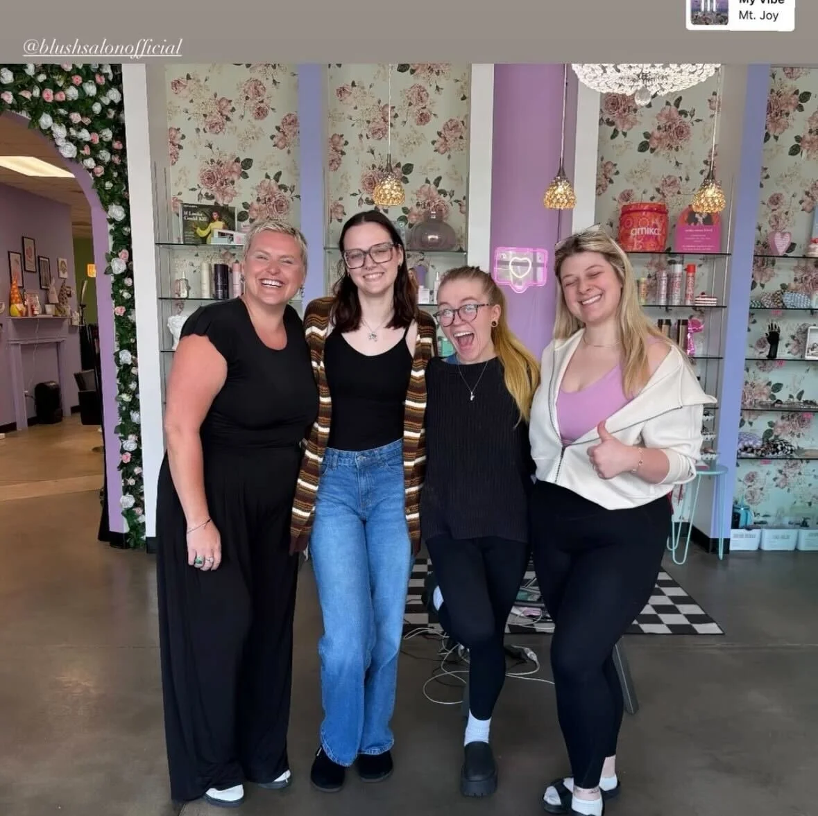 Four women smiling and posing indoors in front of floral wallpaper and decorative shelves.