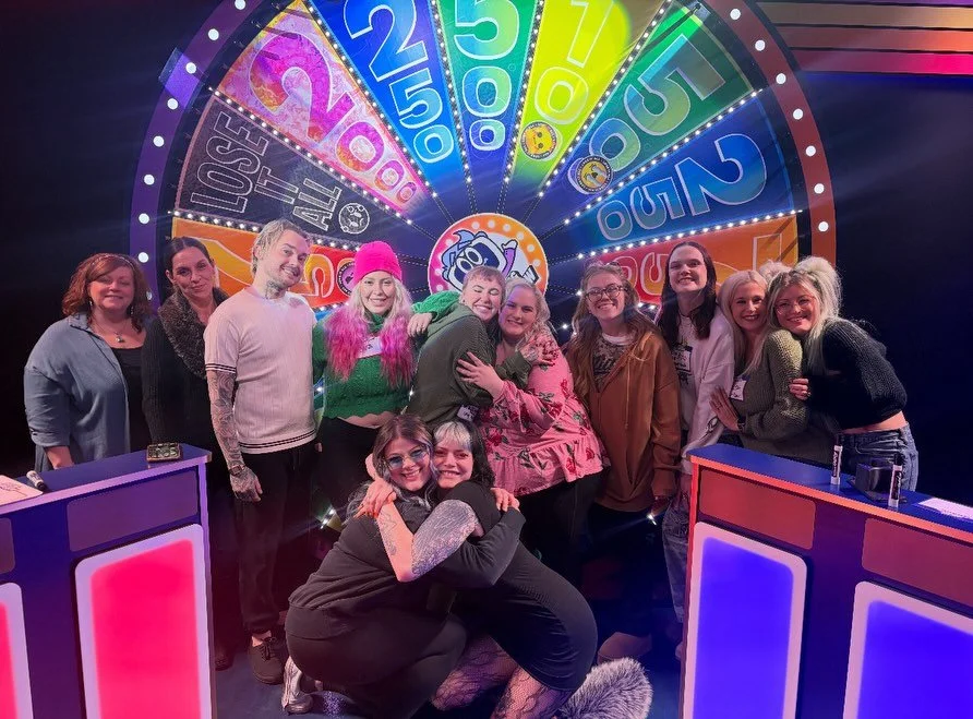 Group of people smiling and hugging in front of a colorful prize wheel at a game show or carnival.
