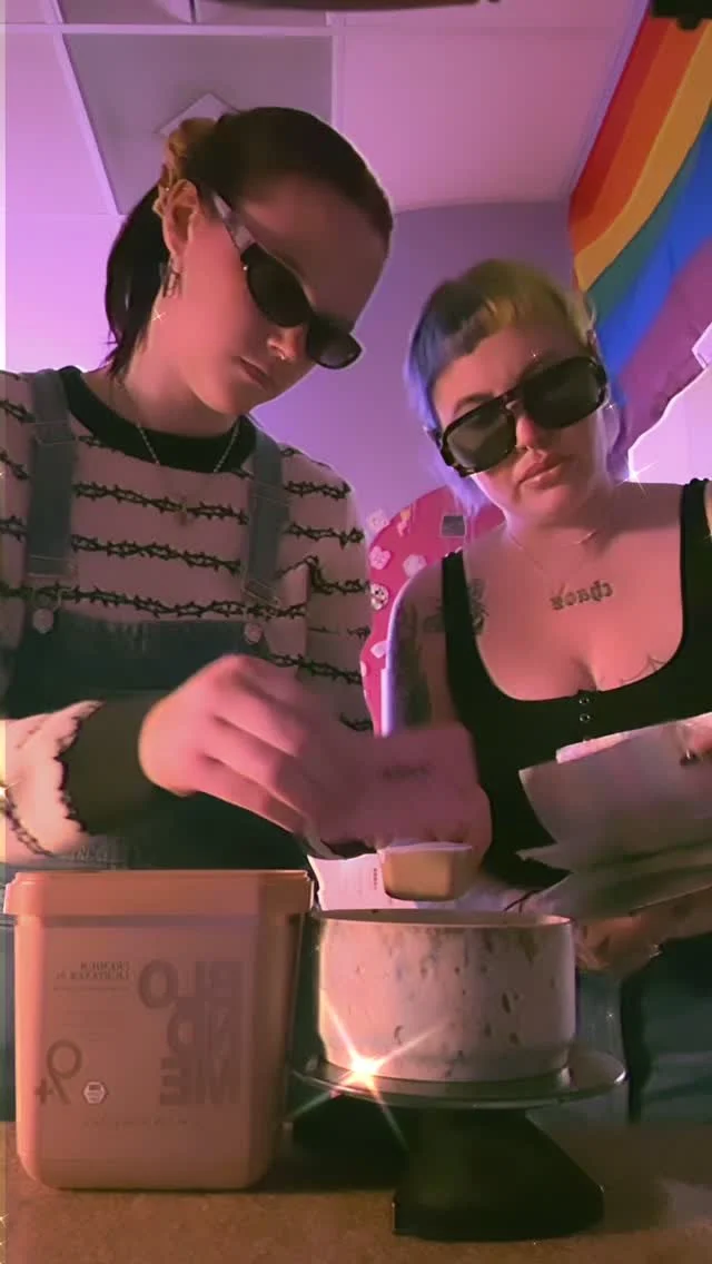 Two women wearing sunglasses at a table with a container marked 'dairy unsweetened' and a marble cake, with rainbow decor in the background.