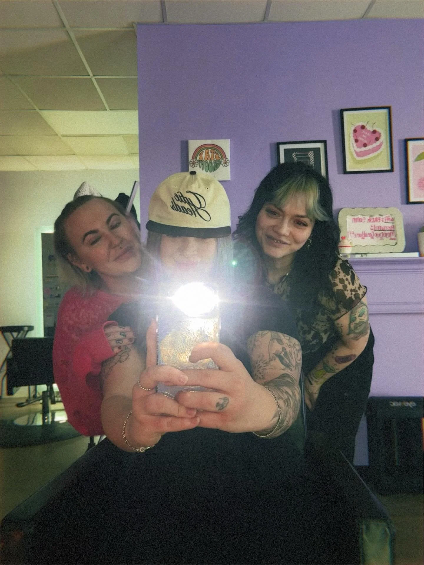 Three women taking a mirror selfie in a room with purple walls, with tattoos on their arms, and frames of art and a cake sign behind them.
