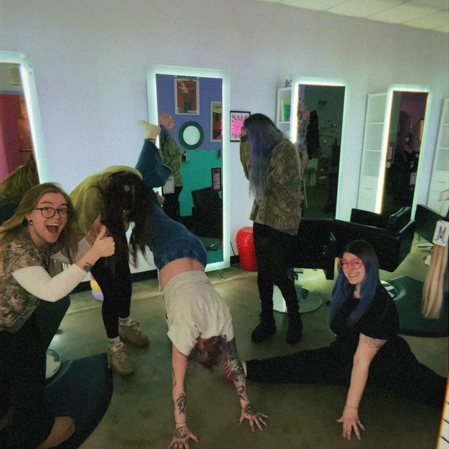 Group of five women having fun in a salon with luminous mirrors, one doing a handstand, one giving a thumbs-up, and others smiling or standing.
