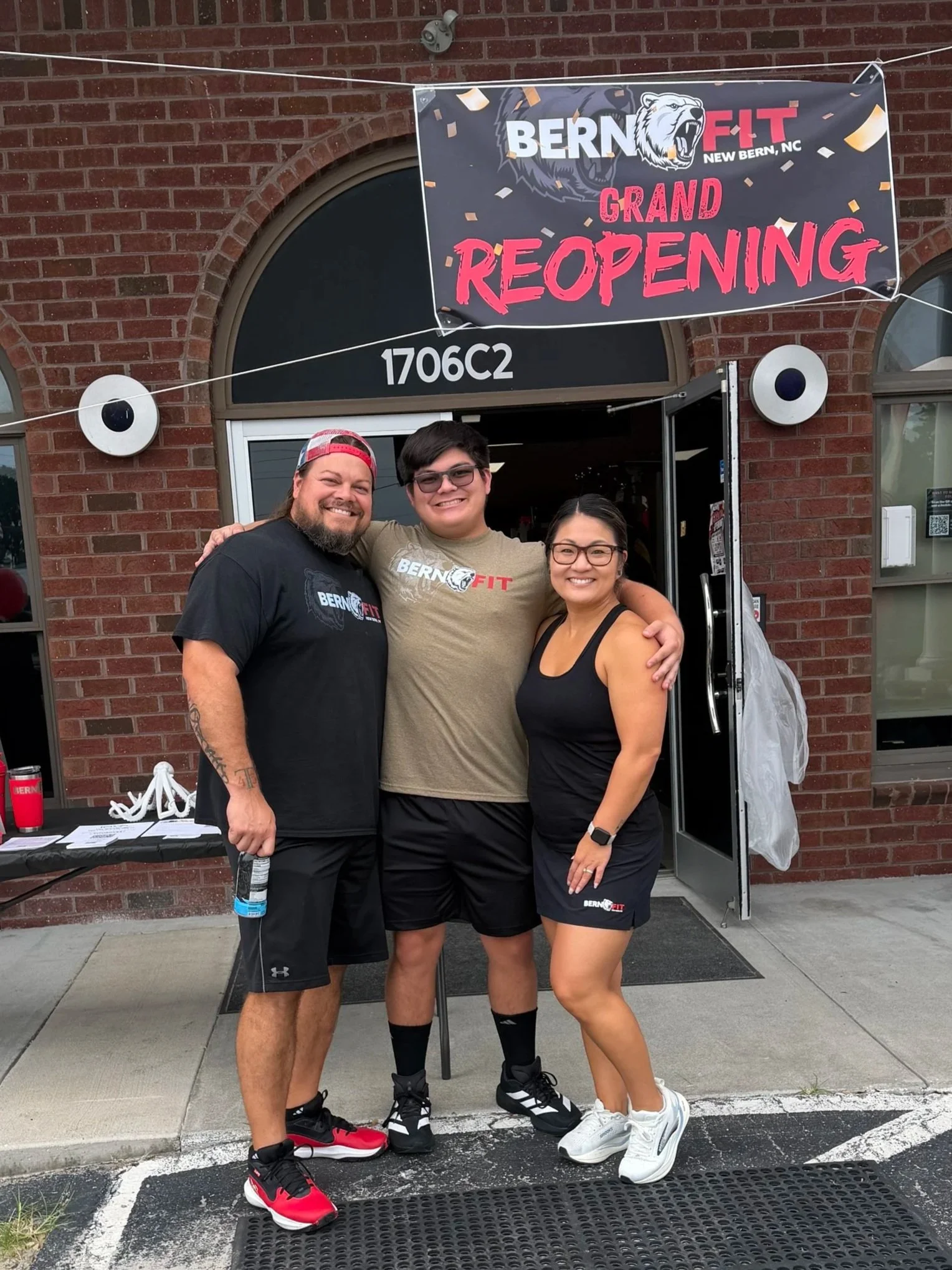 Bern Fit owners Ashley, Kia and Anne Wood outside the gym entrance under a grand reopening banner in New Bern, NC