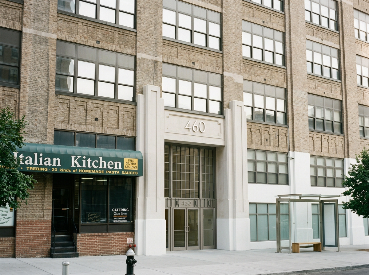 Facade of a multi-story building with large windows, a green awning for Italian Kitchen restaurant, and a glass bus shelter with a wooden bench outside.