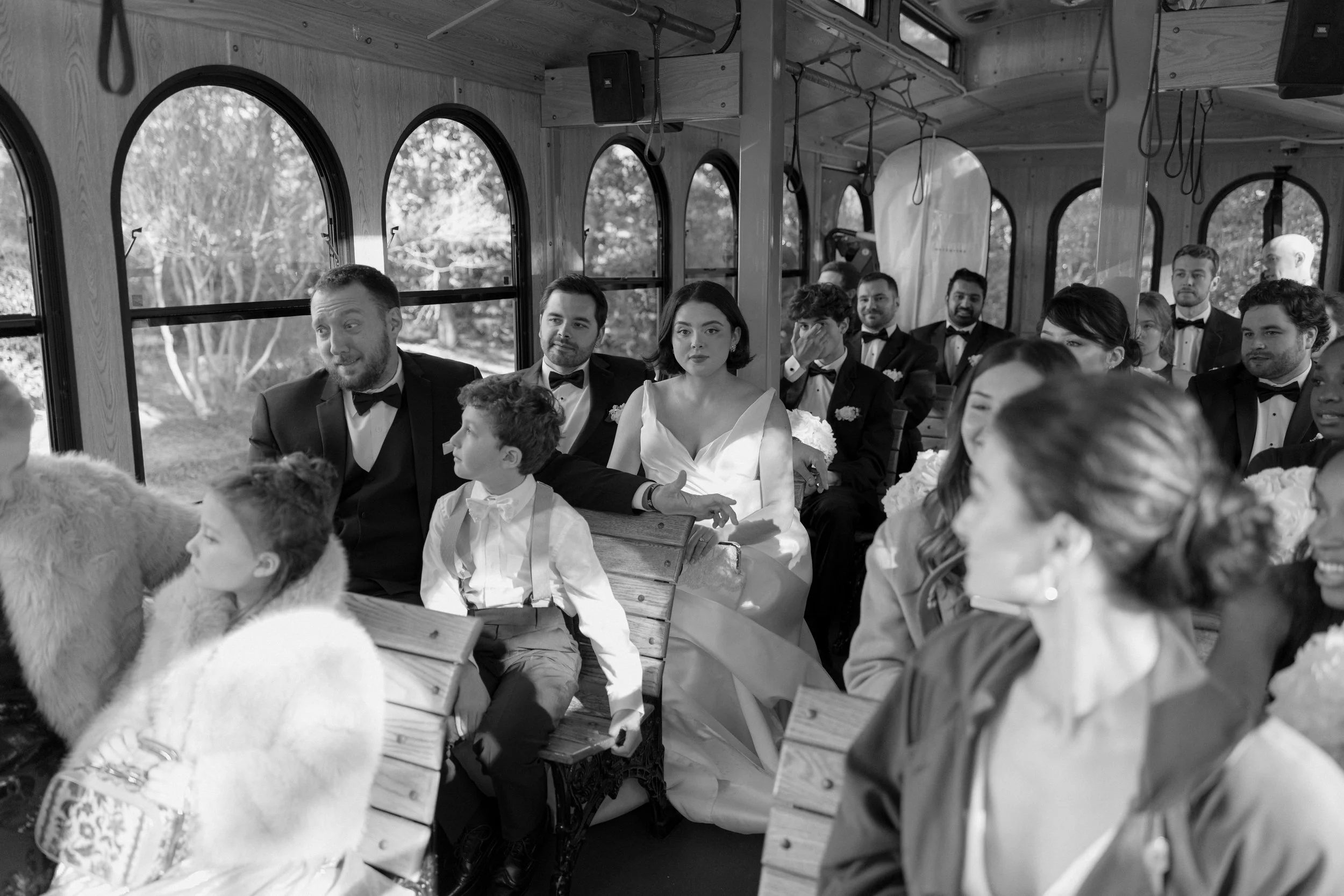 Wedding guests seated inside a trolley bus, dressed in formal attire, with some wearing tuxedos and a bride in a wedding gown.