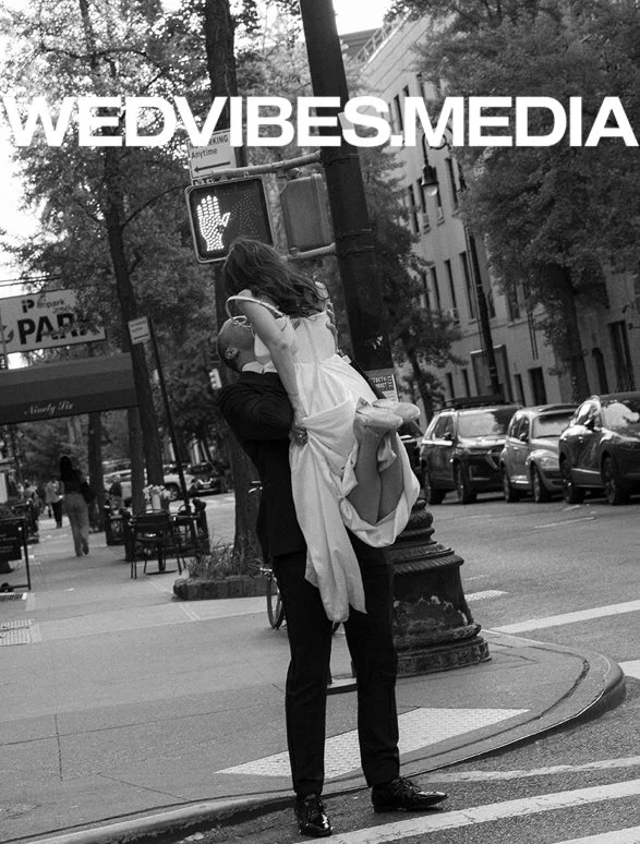 A man in formal attire holding a woman in a wedding dress in an urban street scene with parked cars, trees, and a pedestrian crossing signal.