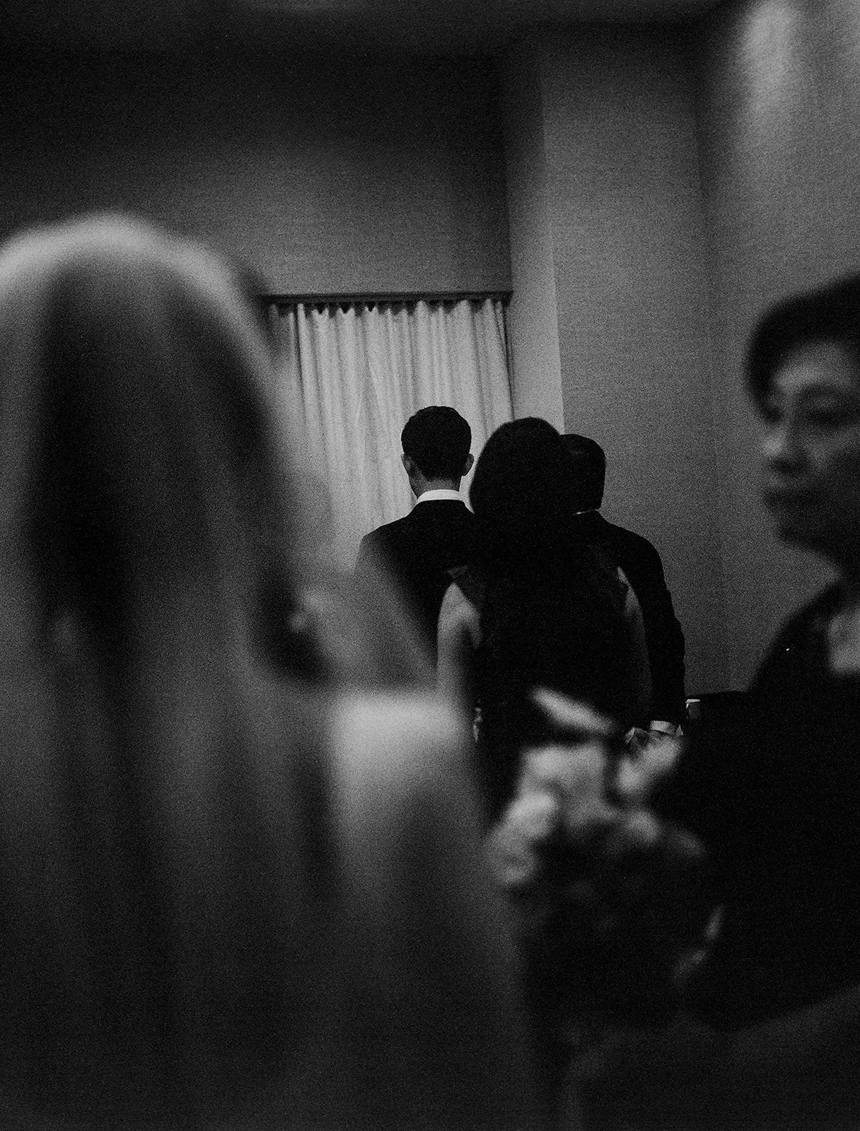 Black and white photo of people at a formal gathering, with a man in a suit standing at the back near a curtain.
