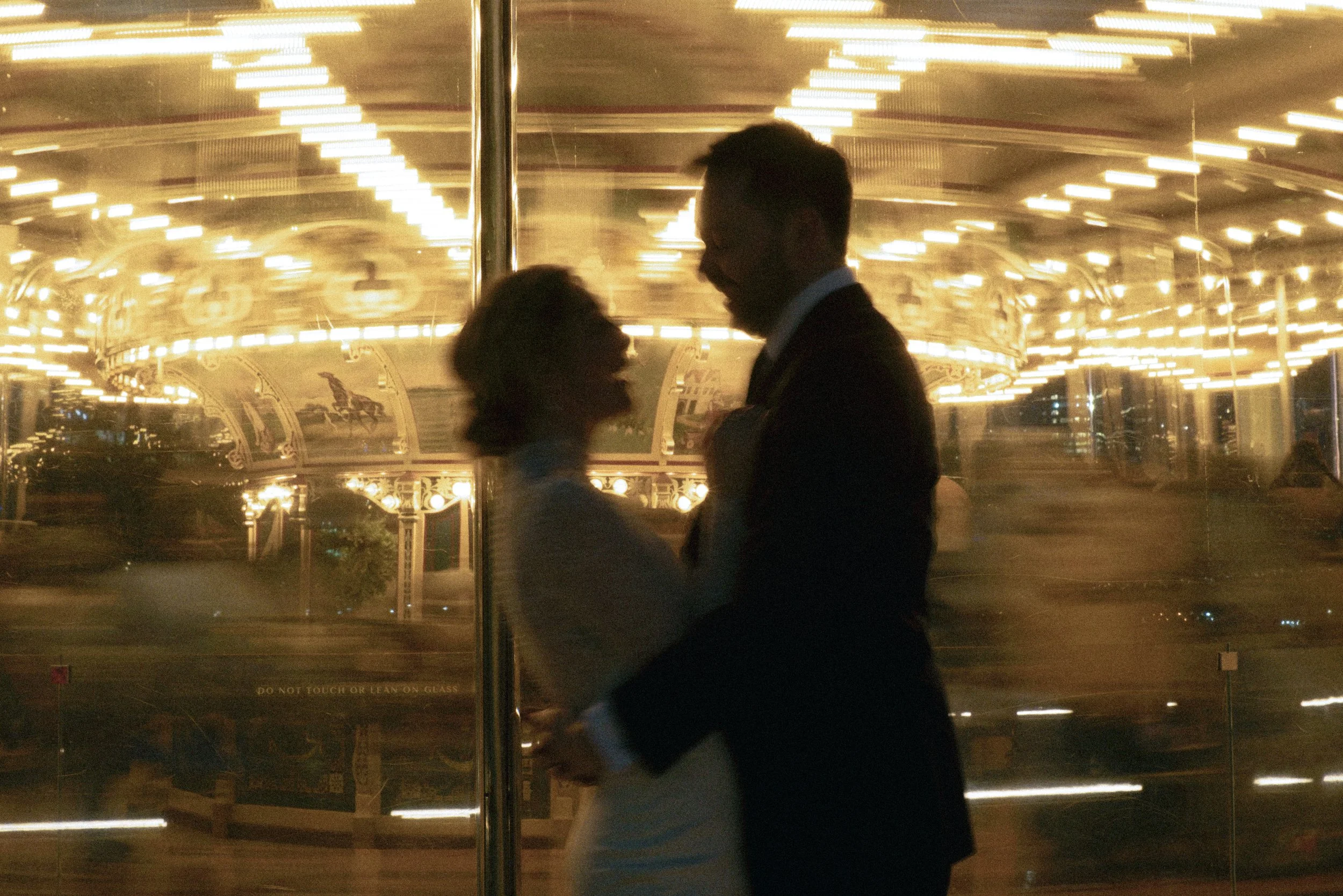 Silhouetted couple dancing closely, possibly sharing a kiss, in front of illuminated carousel ride at night.