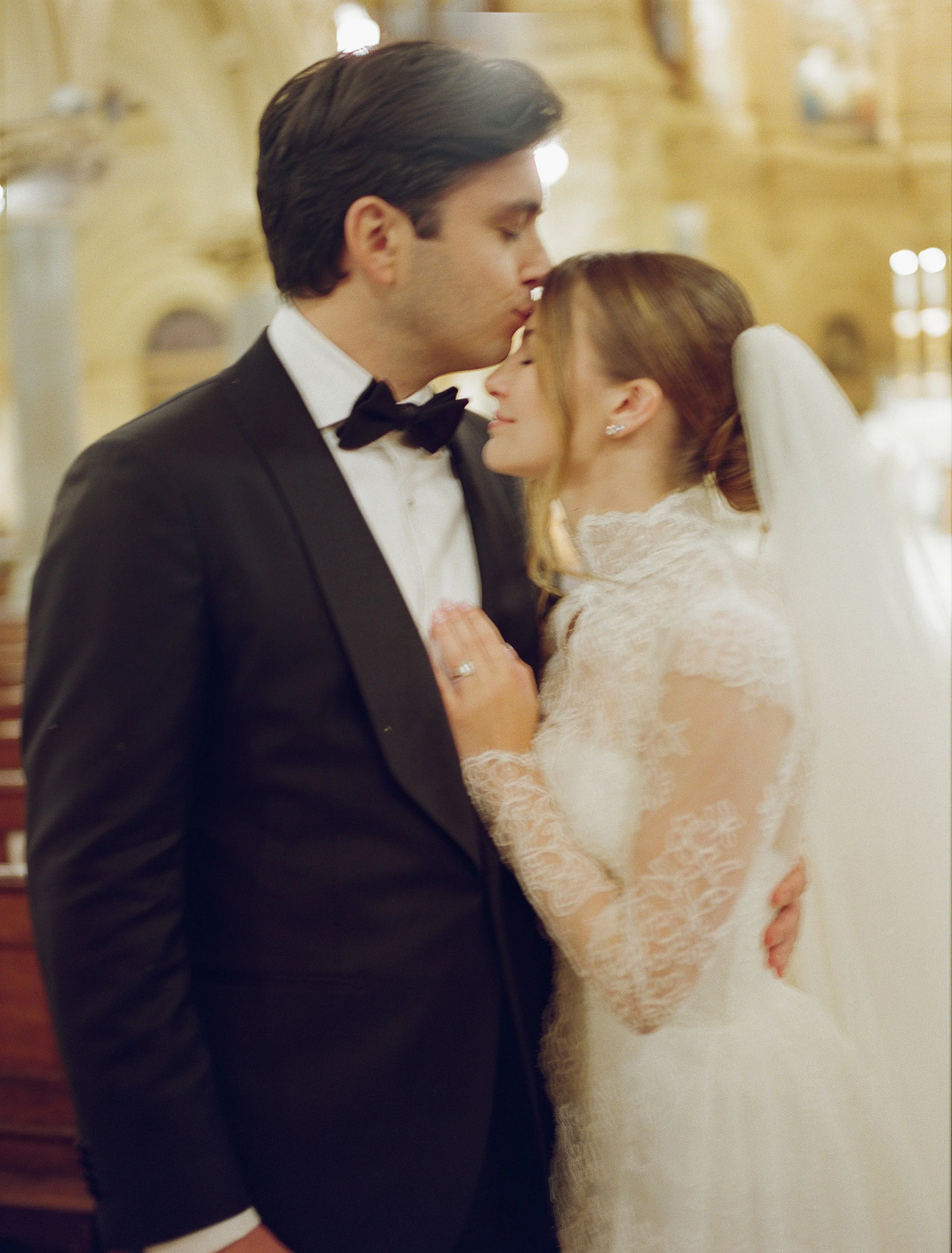 A bride and groom sharing a tender moment during their wedding ceremony indoors. The groom is dressed in a black tuxedo with a bow tie, and the bride is wearing a white lace wedding gown with a veil. The groom is kissing the bride's forehead as she g