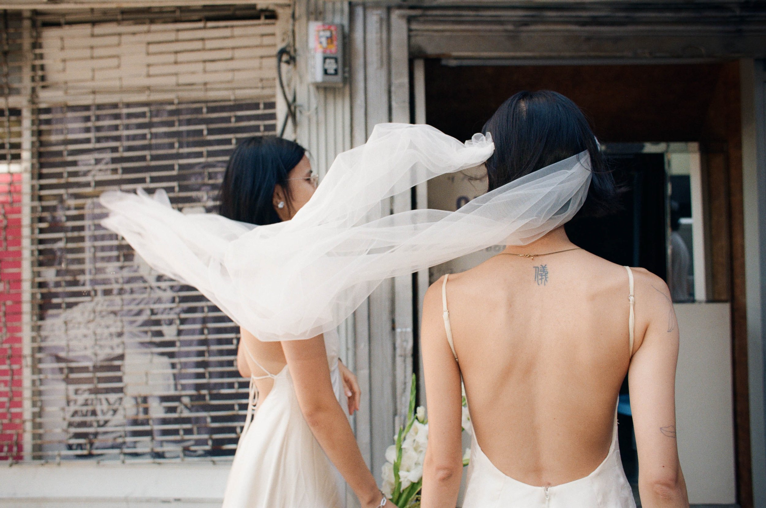 Two women in wedding dresses, one with a veil flying in the wind, standing outside near a building with a metal shutter and brick wall.