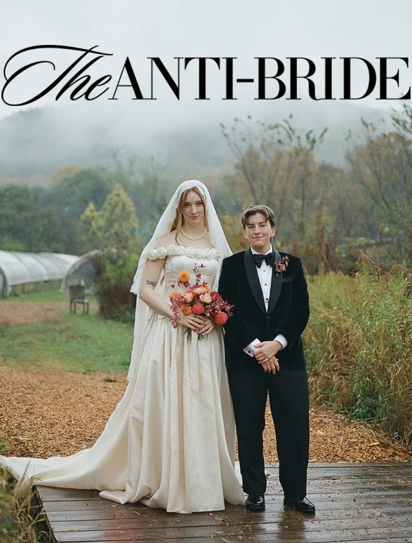 A bride in a vintage wedding dress with a veil holding a flower bouquet and a groom in a tuxedo, standing outdoors on a wooden pathway with trees and greenery in the background.