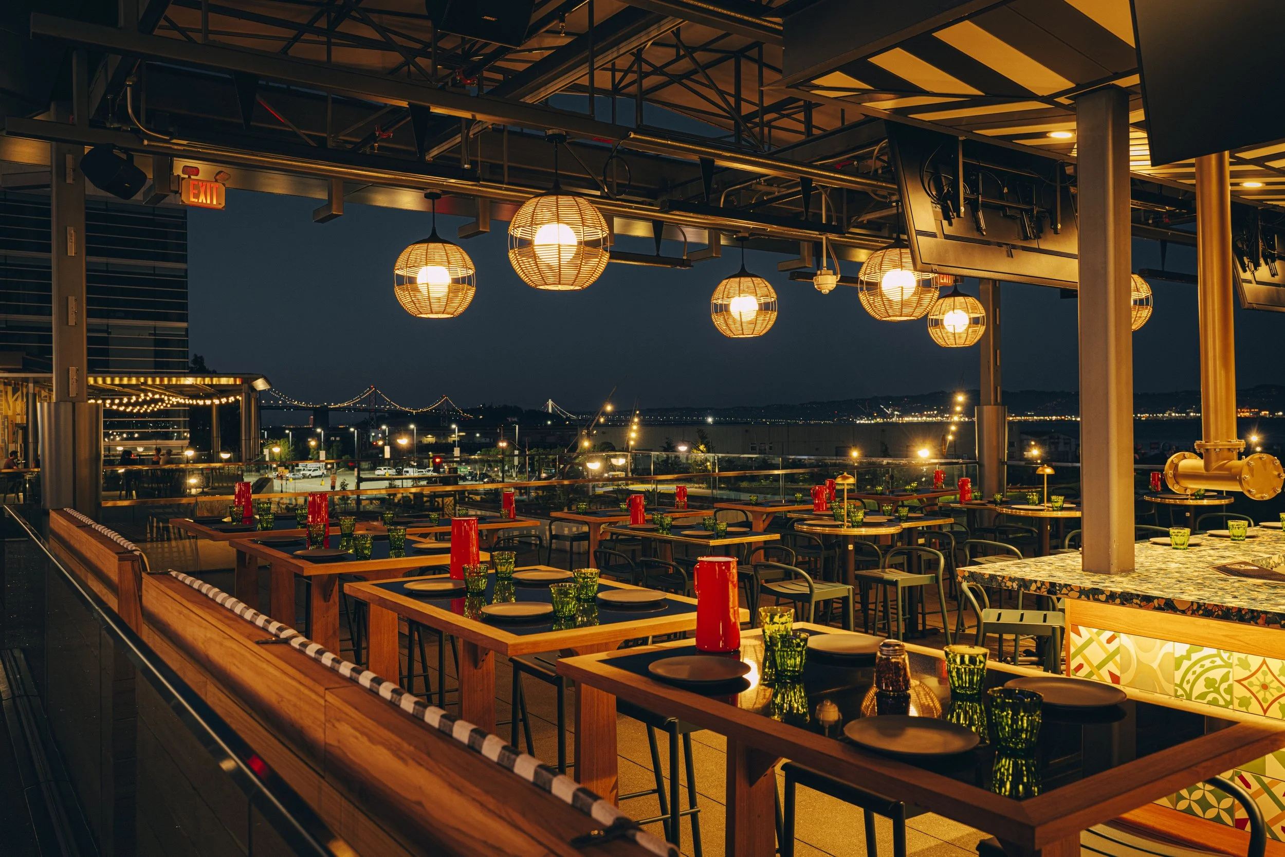 Nighttime scene of the outdoor restaurant and bar with tables set, colorful glassware, and hanging lights, overlooking a city skyline and bridge in the distance.