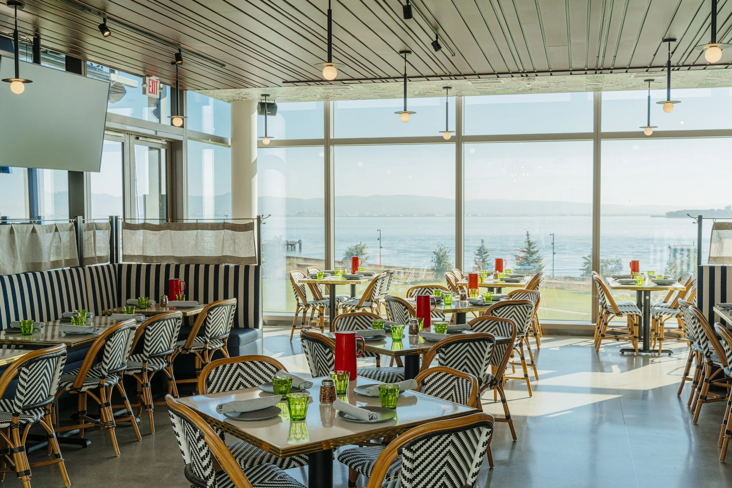 Bright, modern restaurant image with large windows overlooking a body of water and a green landscape, decorated with tables set with green glasses, white napkins, and red condiment bottles.