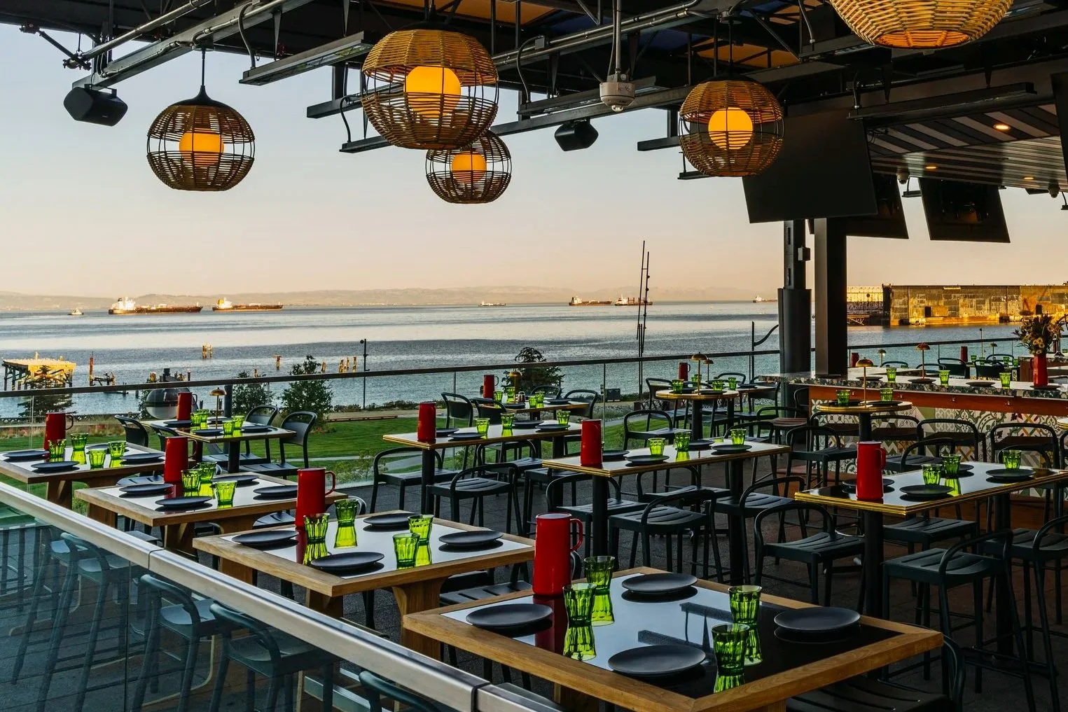 Outdoor restaurant seating with black tables, green glasses, red bottles, and black chairs overlooking a body of water with ships and a distant shoreline during sunset.