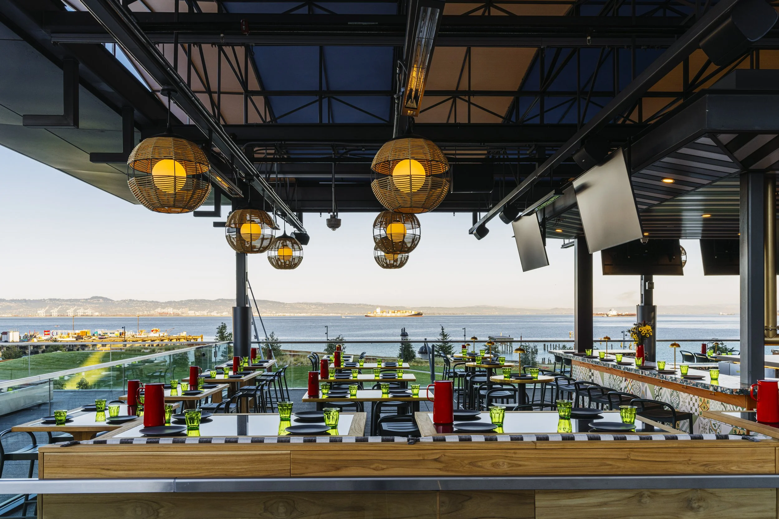 Interior view of the restaurant with tables set with green glasses, black plates, red condiment bottles, and paper napkins, overlooking a body of water with ships and distant land, decorated with hanging wicker lamps.