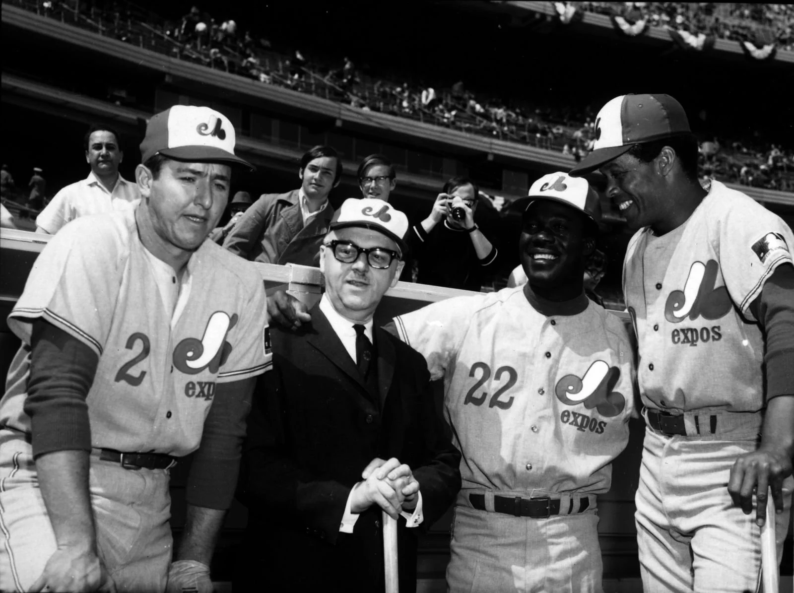 Black and white photograph of four baseball players in uniform and one man in a suit on a baseball field, with spectators in the stands behind them. The players are wearing caps with a logo, and their uniforms have numbers and the word 'expos' on them. They are smiling and appear to be in conversation.