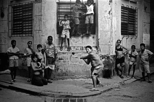 Children gathering outside an old building, some playing with a ball, others watching, in black and white.