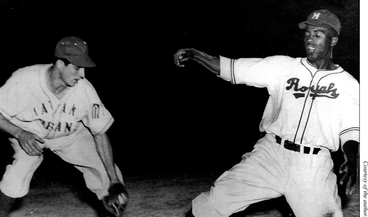 A black and white photograph of two baseball players; one in a Kansas City Monarchs uniform and the other in a Rochester uniform. The player in the Monarchs uniform is reaching down to pick up a baseball from the ground while the other player appears to be preparing to swing a bat.