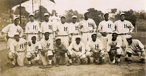 A vintage black-and-white photograph of a baseball team with 14 men wearing uniforms with the letter 'H' on the chest, posed outdoors on a baseball field.