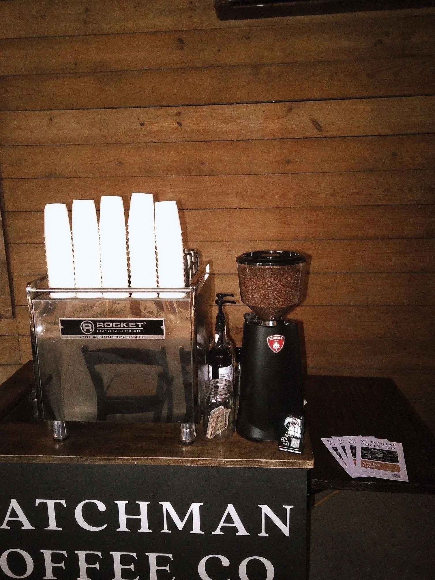 Coffee and espresso station with a Rocket espresso machine, paper cups, a coffee grinder, and sample cards on a wooden counter.
