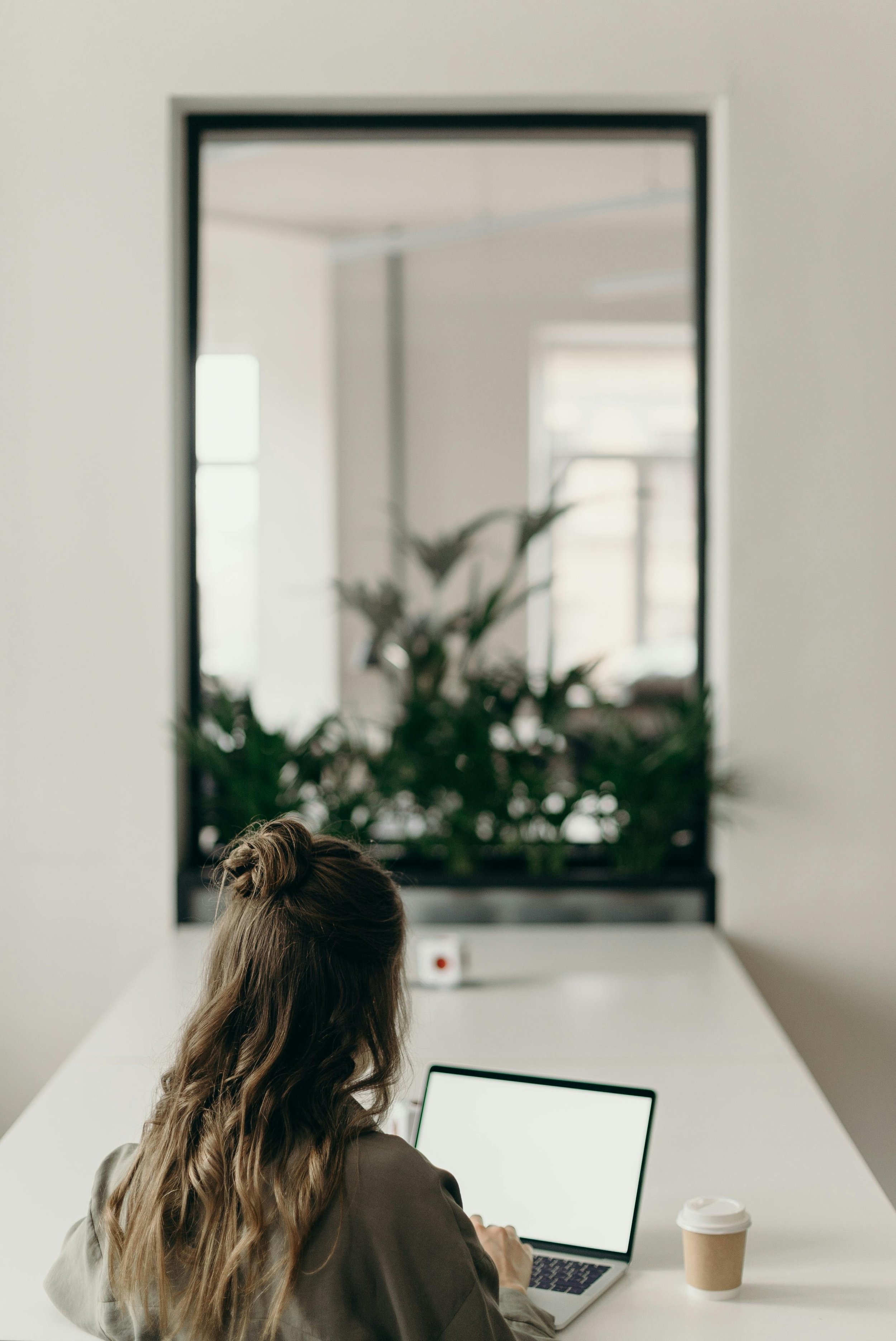 Person using a laptop at a desk with a coffee cup, facing a large window with plants outside.
