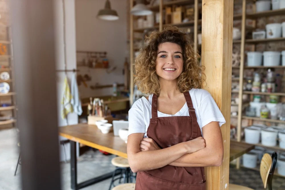 A woman with curly hair wearing a white t-shirt and brown apron standing in a pottery studio, smiling with arms crossed.